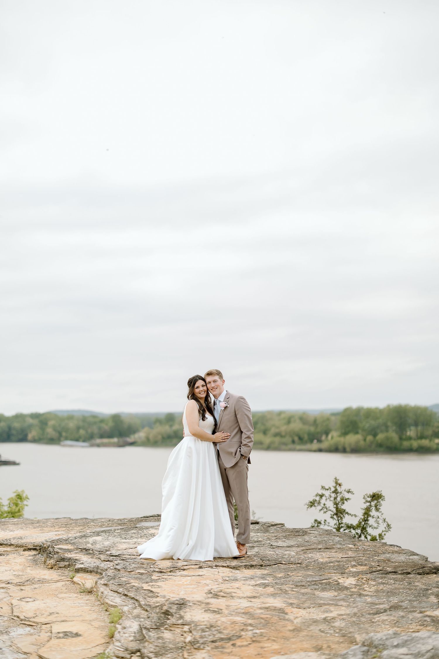 A couple stands on a rocky outcrop overlooking a serene lake surrounded by green trees on an overcast day.