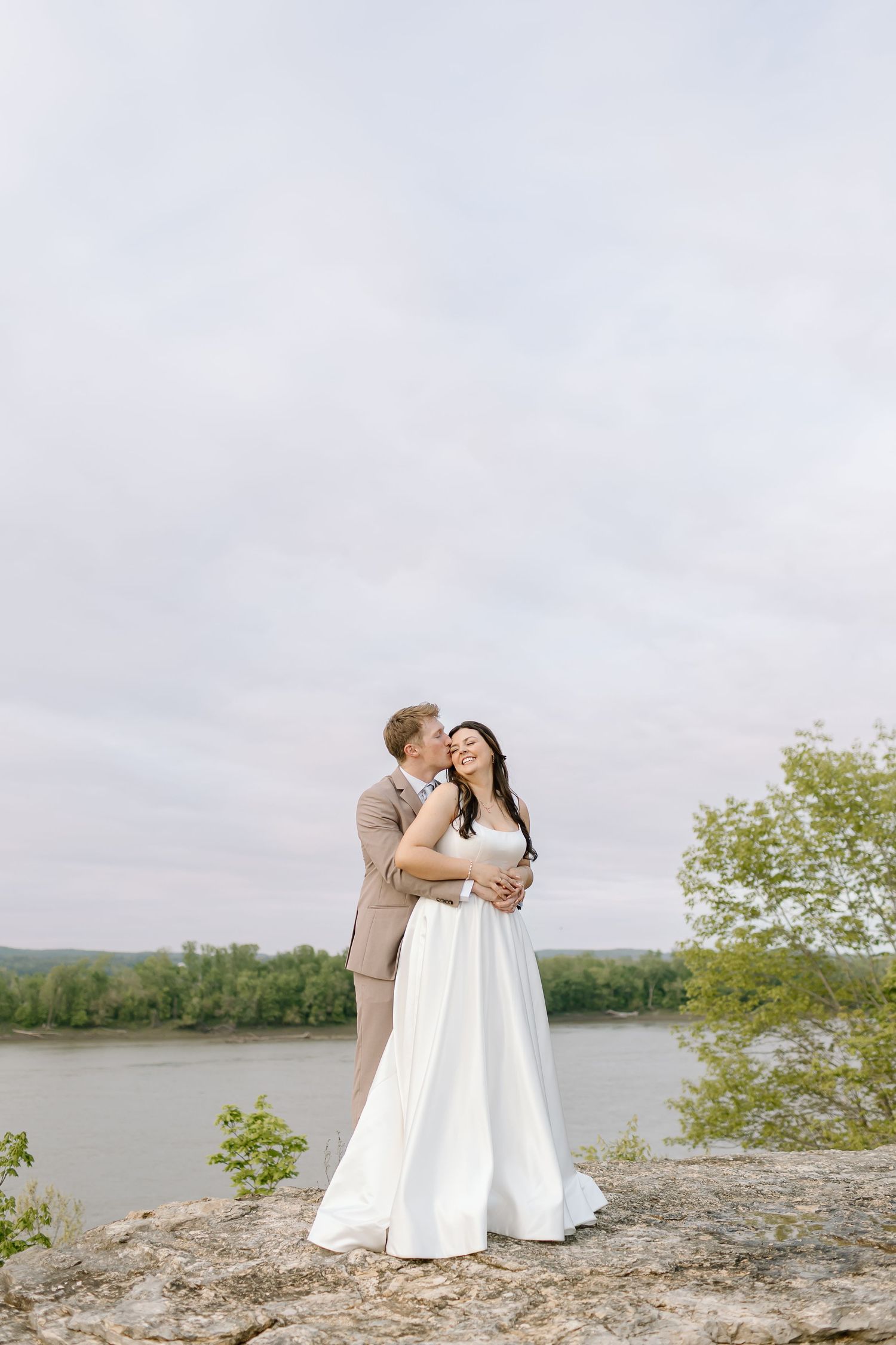 A couple in wedding attire shares romantic moments on a rocky cliff overlooking a serene lake on their special day.