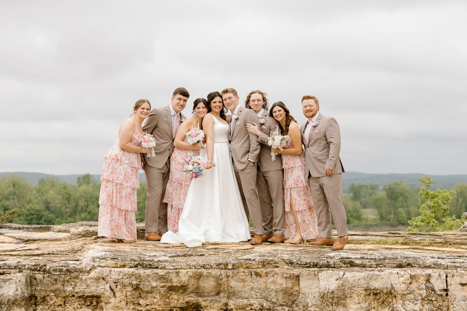 Wedding party in blush pink and tan attire poses together on stone overlook with scenic mountain view in background.