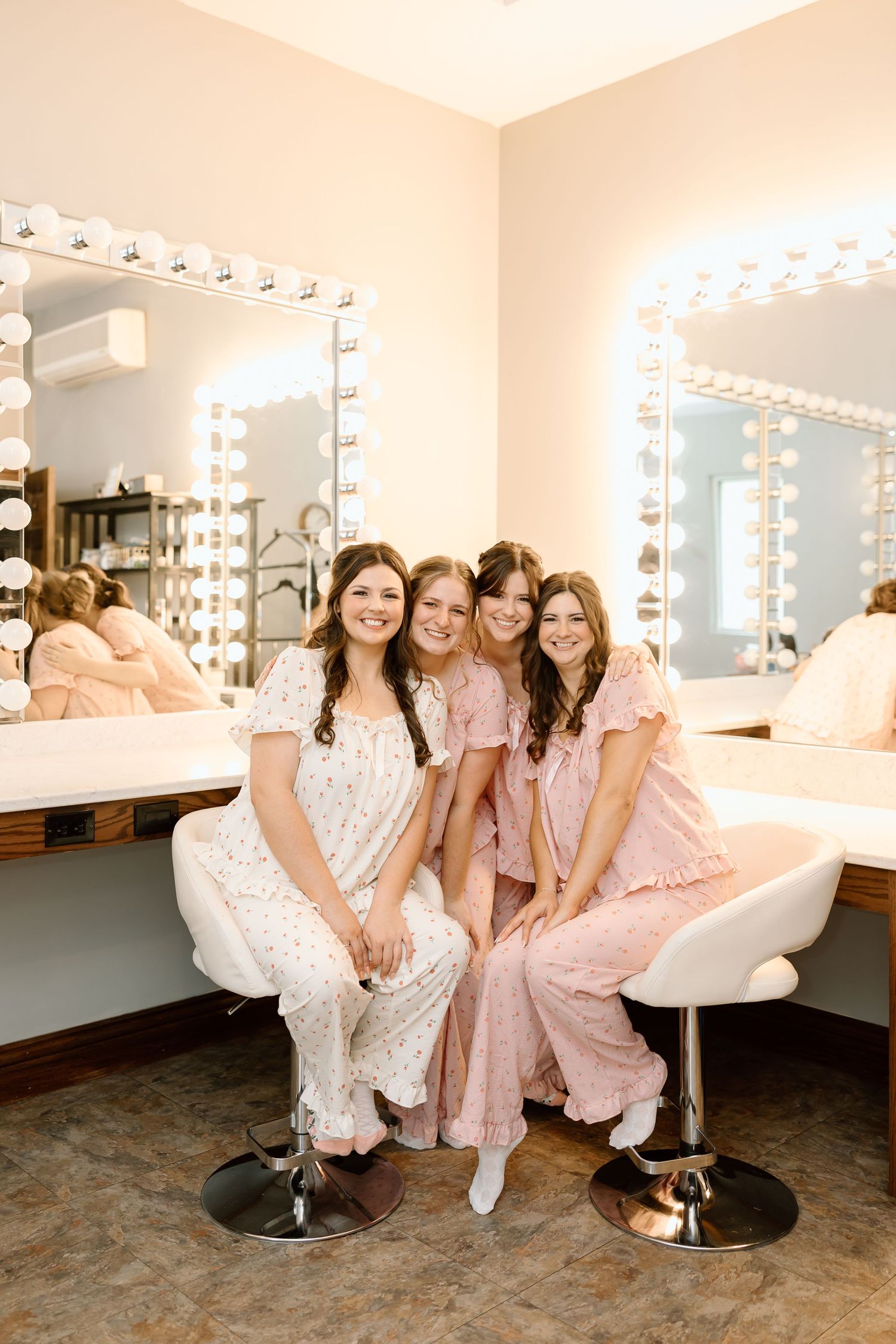 A group poses together in a dressing room with illuminated vanity mirrors and salon chairs in soft pink lighting.