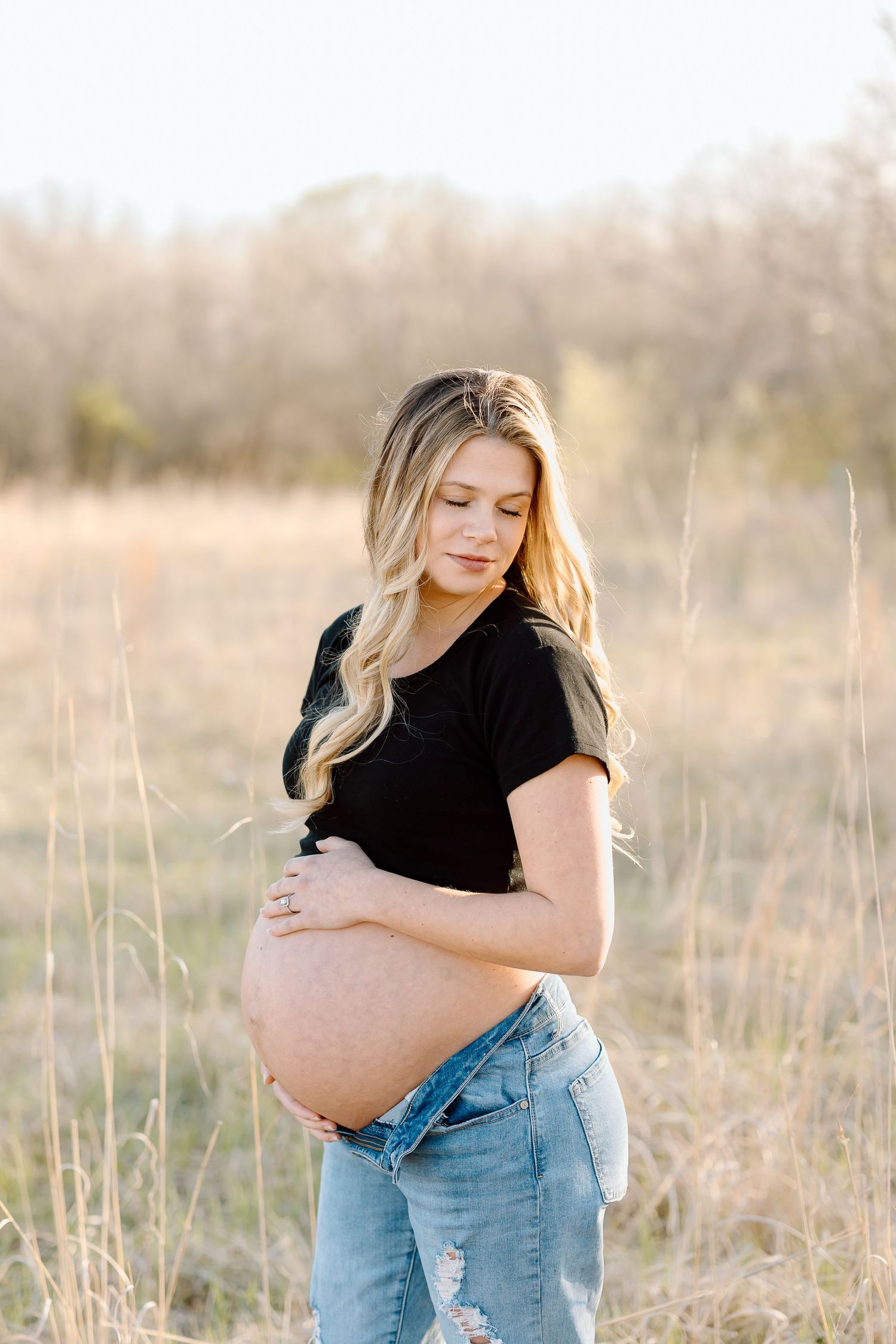 Pregnant woman in casual attire holds her baby bump while posing in a golden-hour field setting.