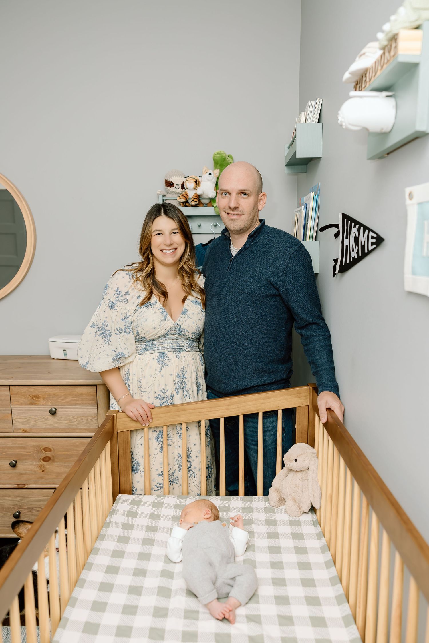 A family poses together in a nursery with a wooden crib and gray walls.