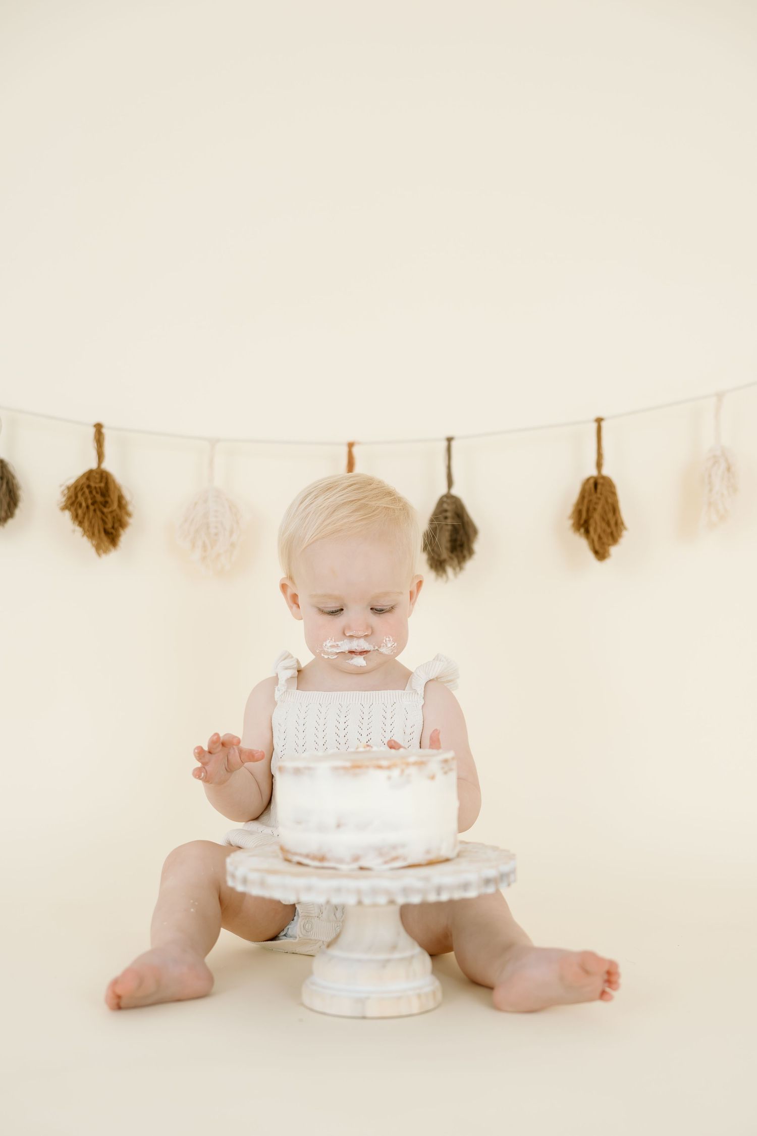 A baby in white sits on a cream background with a decorated birthday cake for a minimalist cake smash photo session.