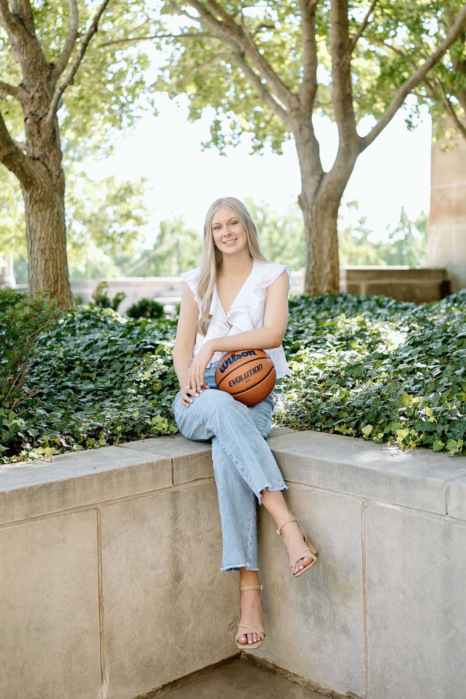 Person in light colored top and jeans sits casually on concrete wall surrounded by lush greenery in outdoor setting.