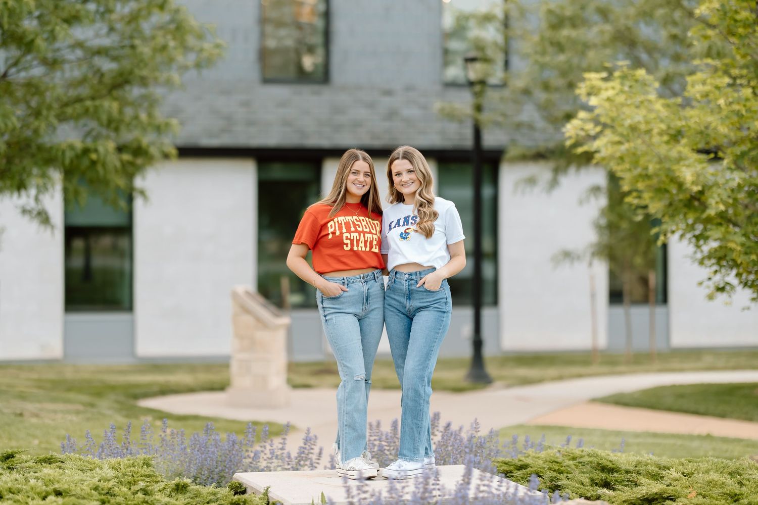 Two people in denim jeans and casual tops stand together on a pathway near white buildings and green trees.