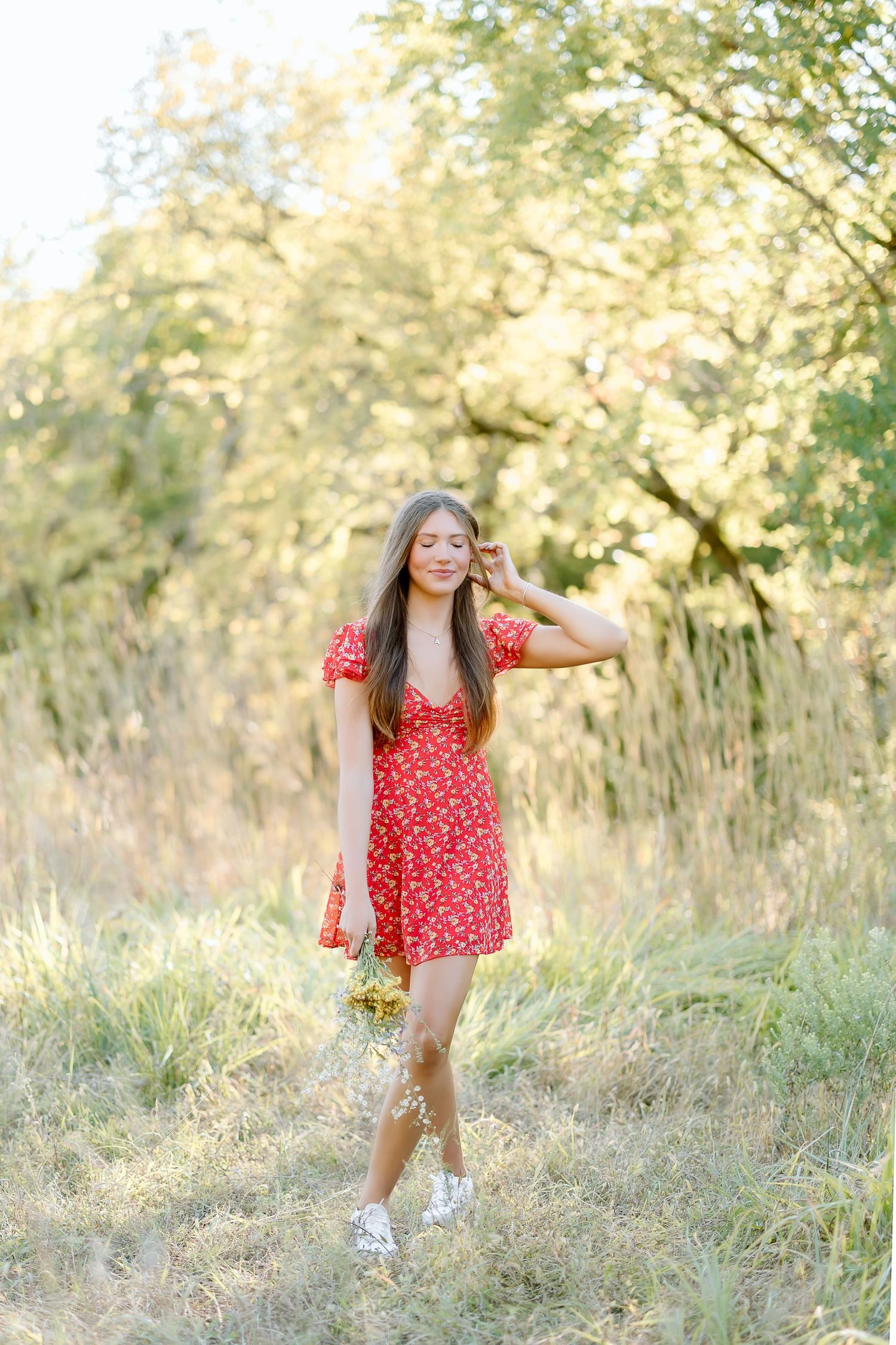 A person in a red floral dress poses playfully in a sunlit field with golden grasses and trees in the background.