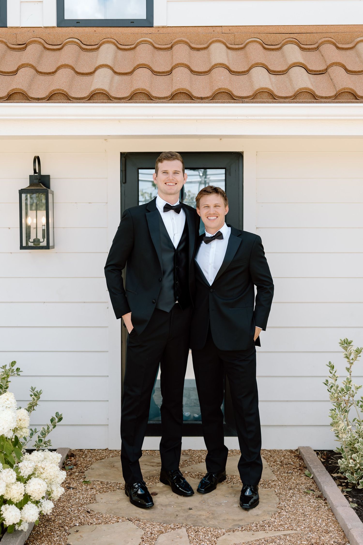 Two grooms in black tuxedos with bow ties smile together outside a white building with terracotta roof tiles.