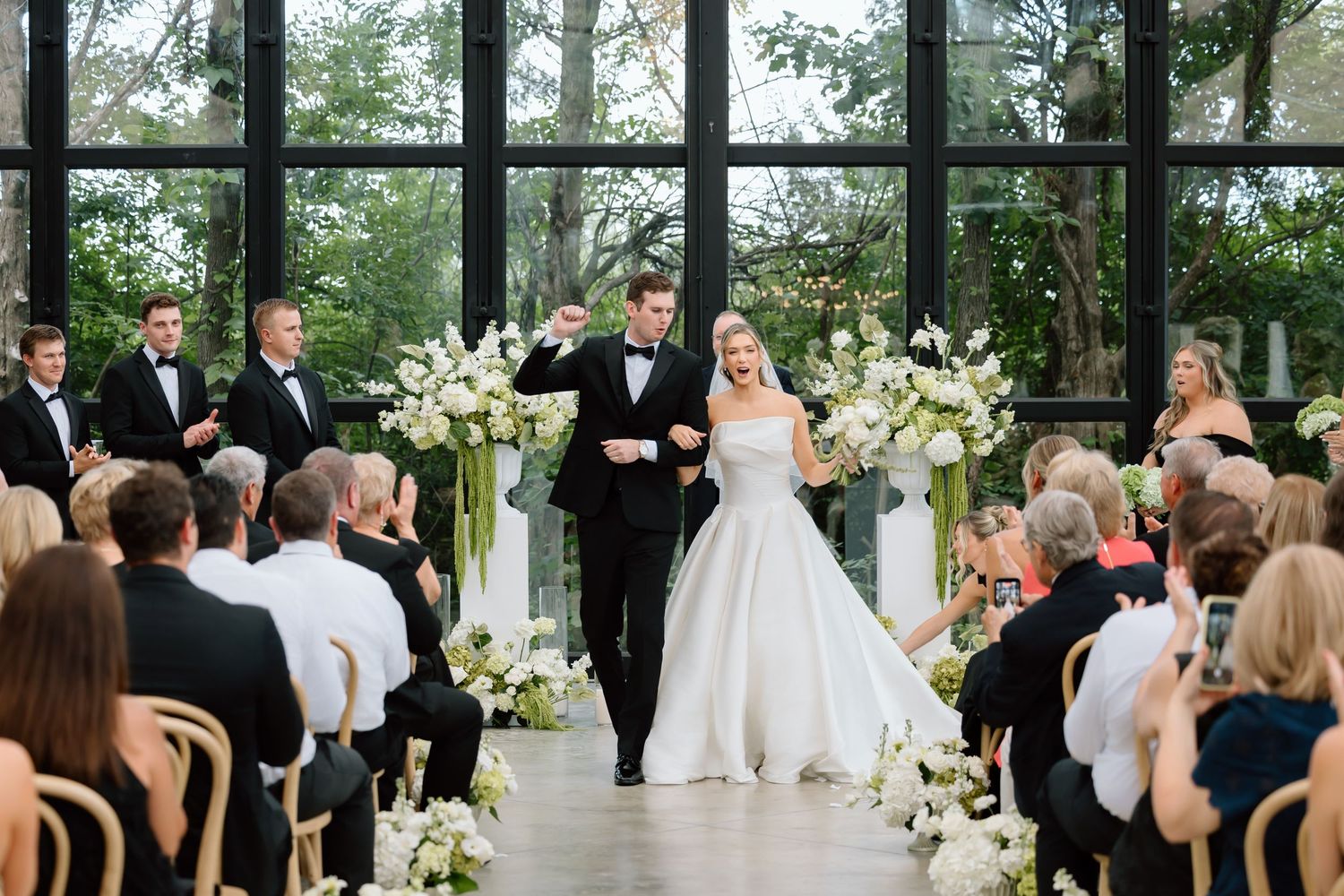 Bride and groom walk down the aisle at elegant wedding ceremony with white floral arrangements and floor-to-ceiling windows.
