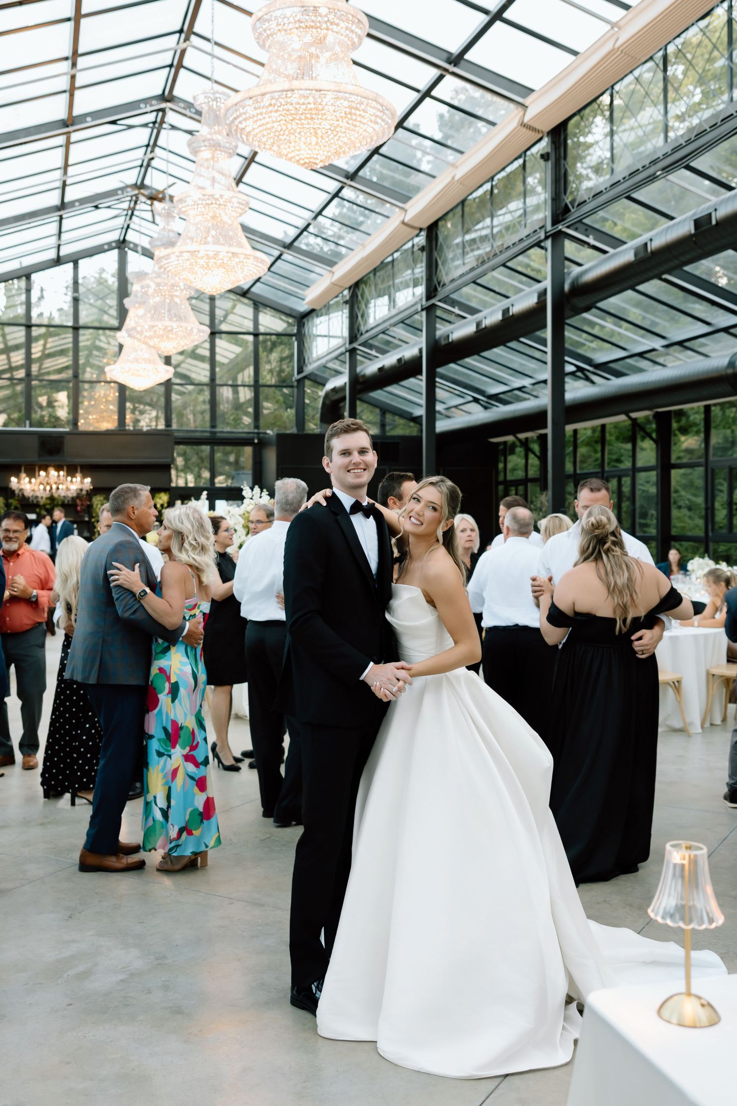 Bride and groom share first dance in elegant glass greenhouse venue with chandeliers and guests dancing behind them.