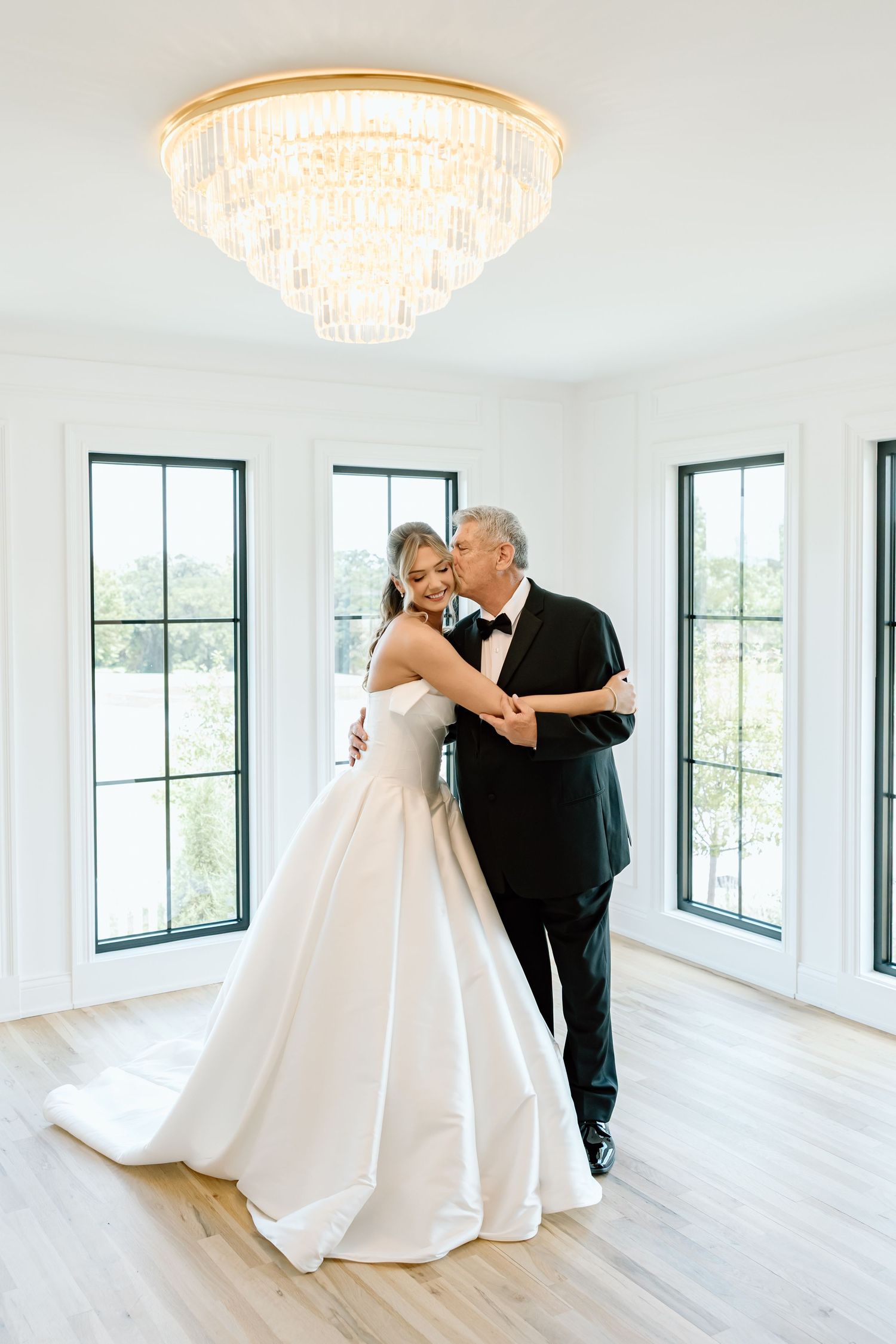 Bride in white ballgown shares a tender father-daughter dance under a crystal chandelier in an elegant white room.