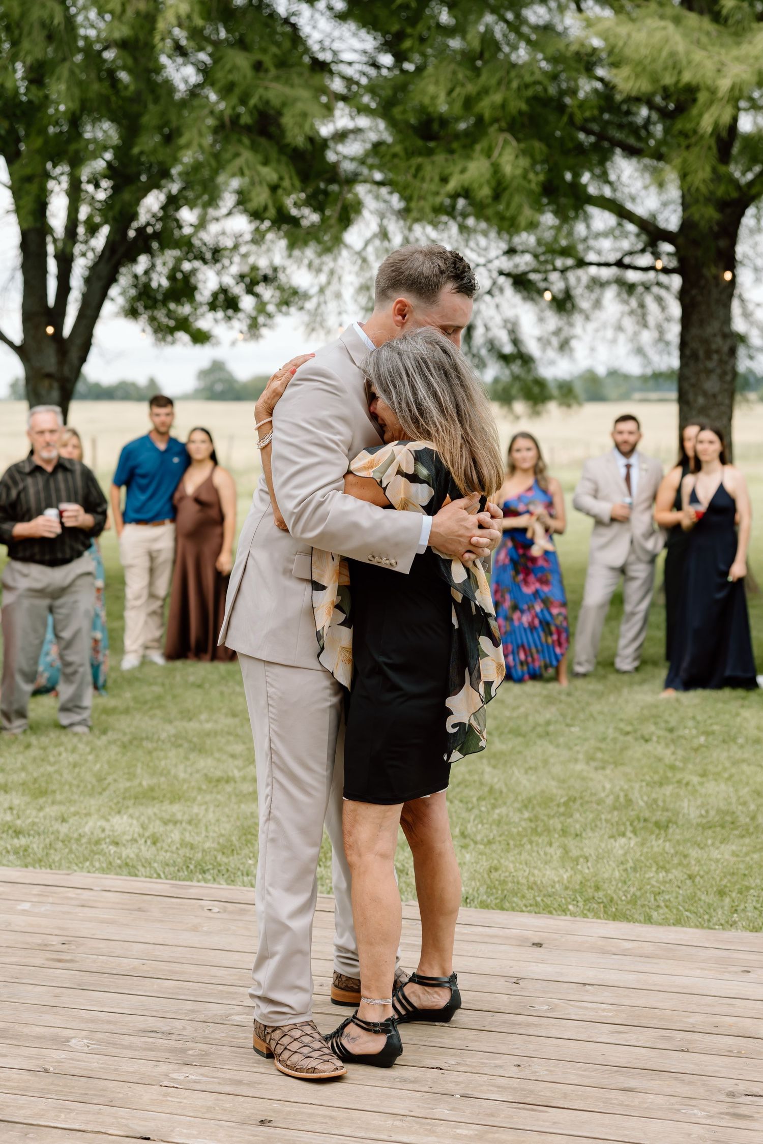 Groom in tan suit shares emotional dance with woman in black dress at outdoor wedding reception on wooden deck.