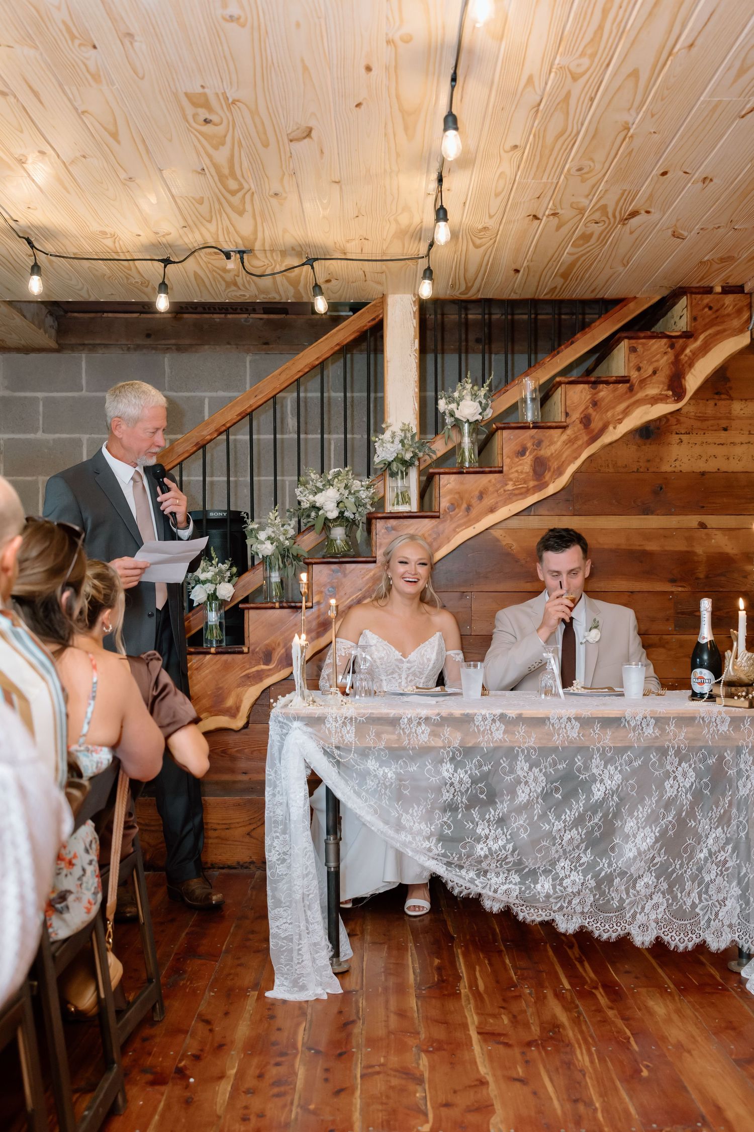 Bride and groom seated at sweetheart table during wedding reception toast in rustic barn venue with string lights.