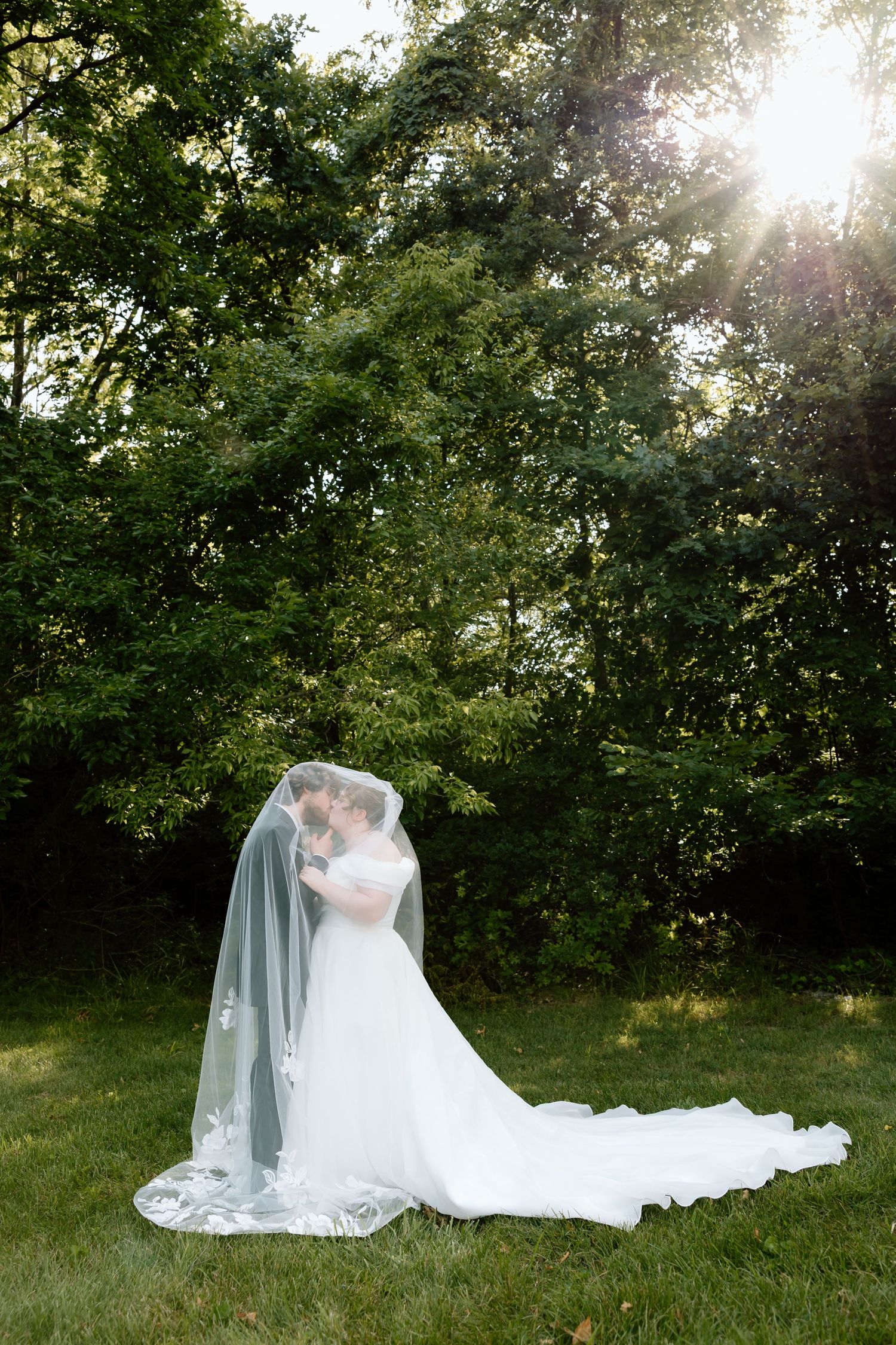 Bride and groom share a kiss under a flowing cathedral veil in a sunlit garden setting.
