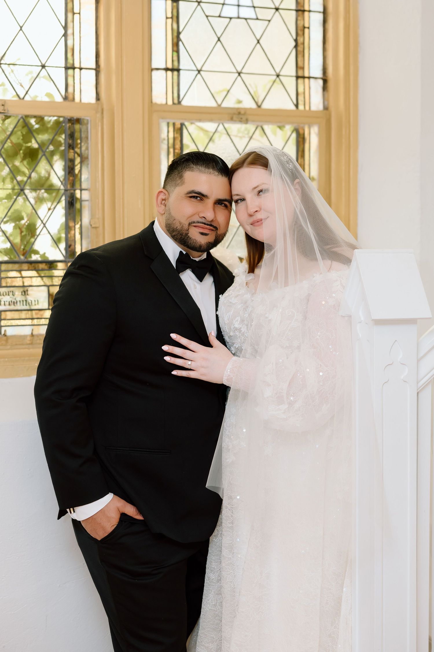 Bride and groom pose elegantly in a chapel, she in a beaded gown with veil, he in a black tuxedo.