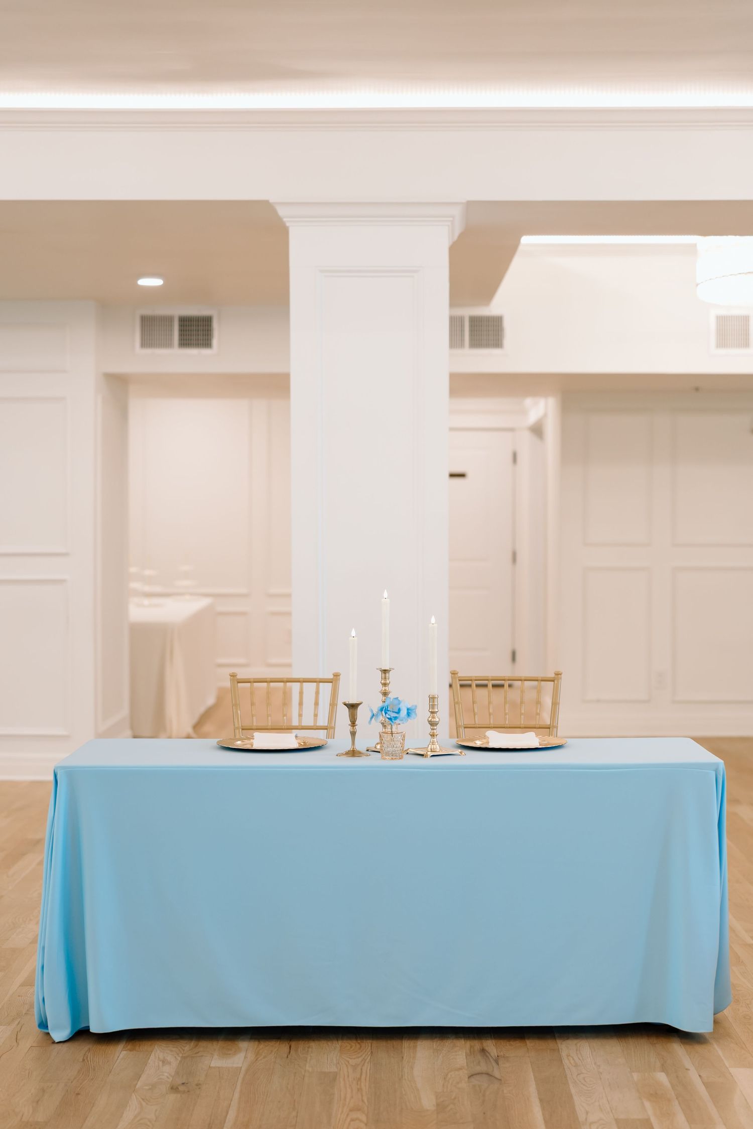 Elegant sweetheart table with light blue linen, gold chairs, white candles, and blue floral centerpiece in a bright venue.