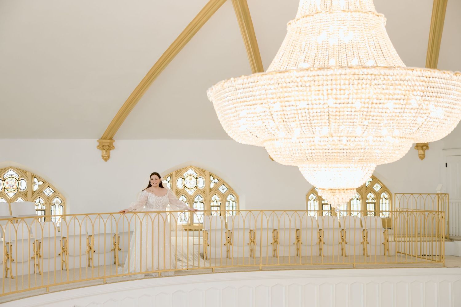 Elegant white and gold ballroom interior with crystal chandelier and woman standing on ornate balcony railing.