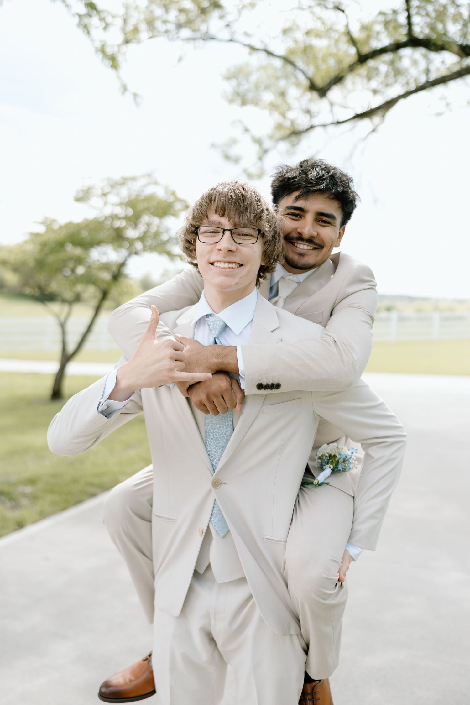 Two young men in matching beige suits embrace outdoors at a formal event, one giving a shaka sign.
