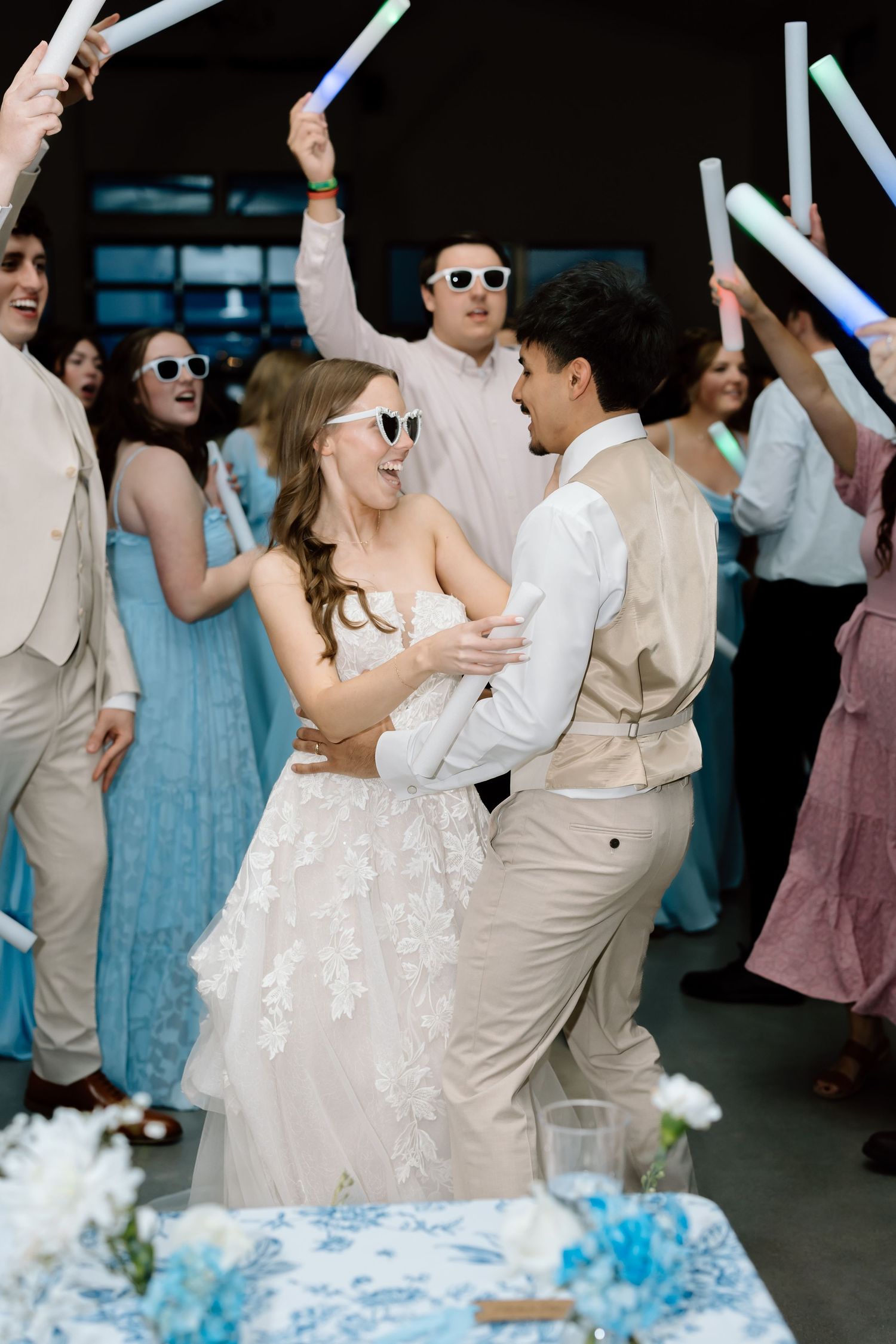 Bride and groom dance joyfully surrounded by guests waving glow sticks at a festive wedding reception.