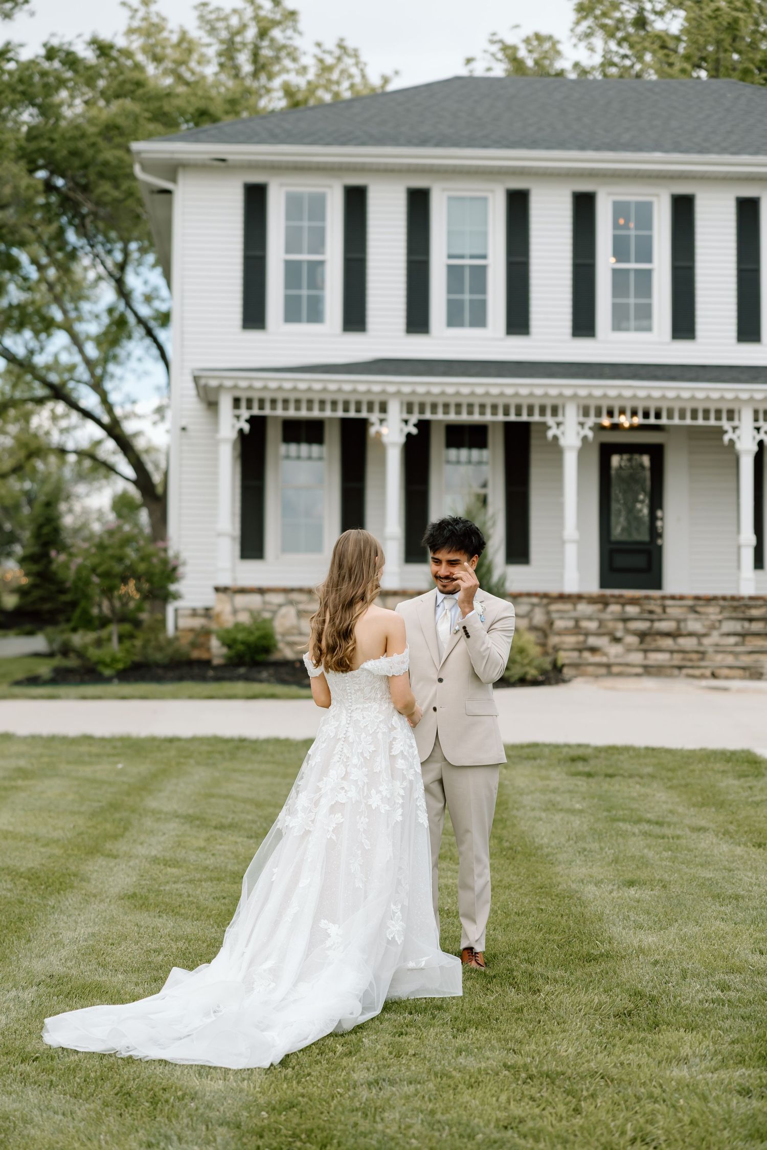 Bride and groom share a first look moment on green lawn before a white two-story farmhouse with dark shutters.