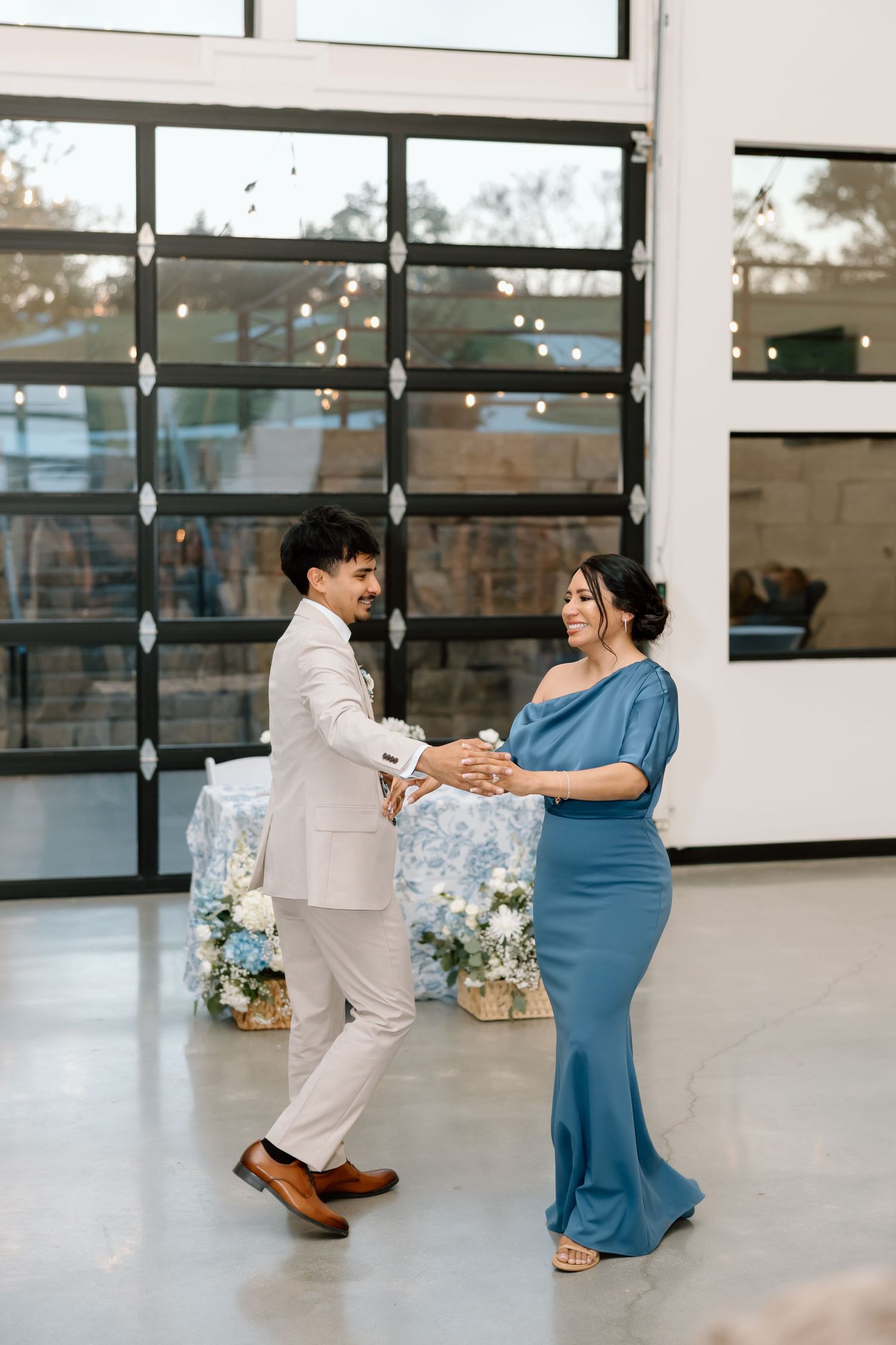 Couple sharing a romantic dance in an elegant event venue with large windows and floral arrangements.