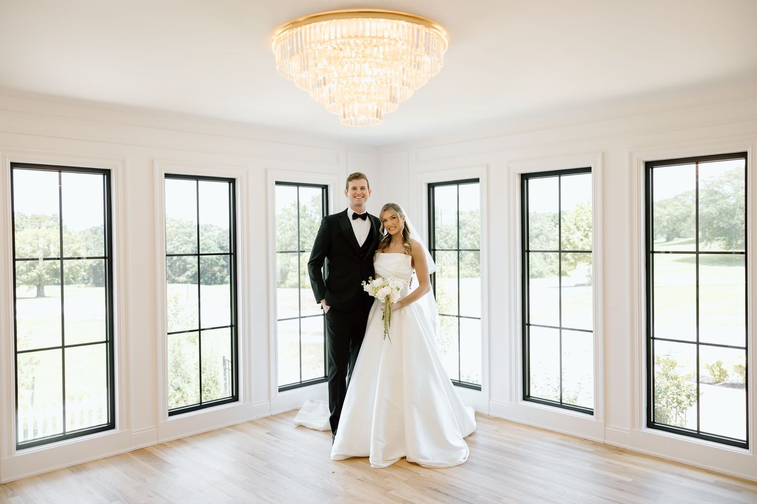 Bride and groom posing elegantly in a bright white room with black-framed windows and a gold chandelier.