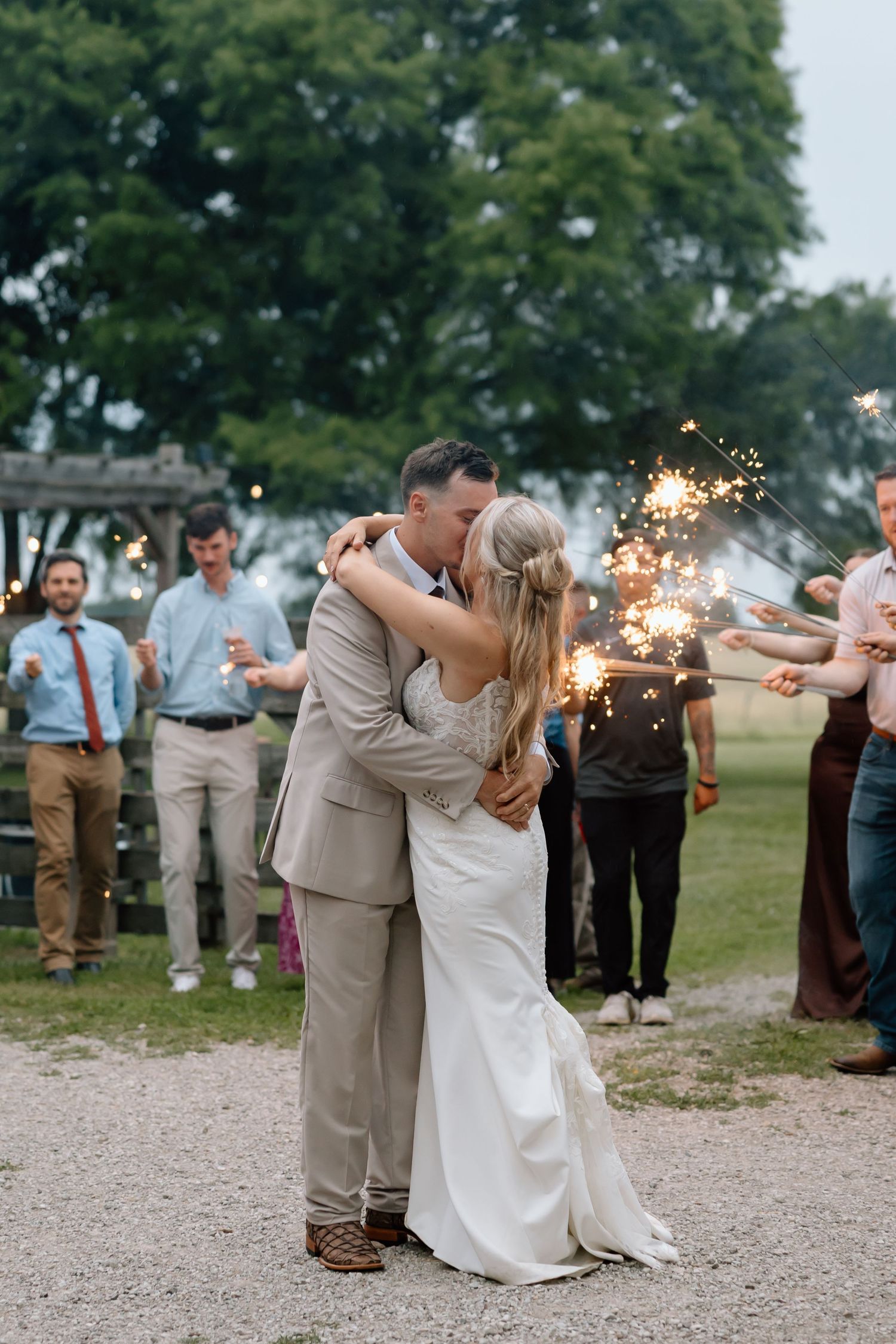 Bride and groom share a kiss surrounded by wedding guests holding sparklers at an outdoor evening celebration.
