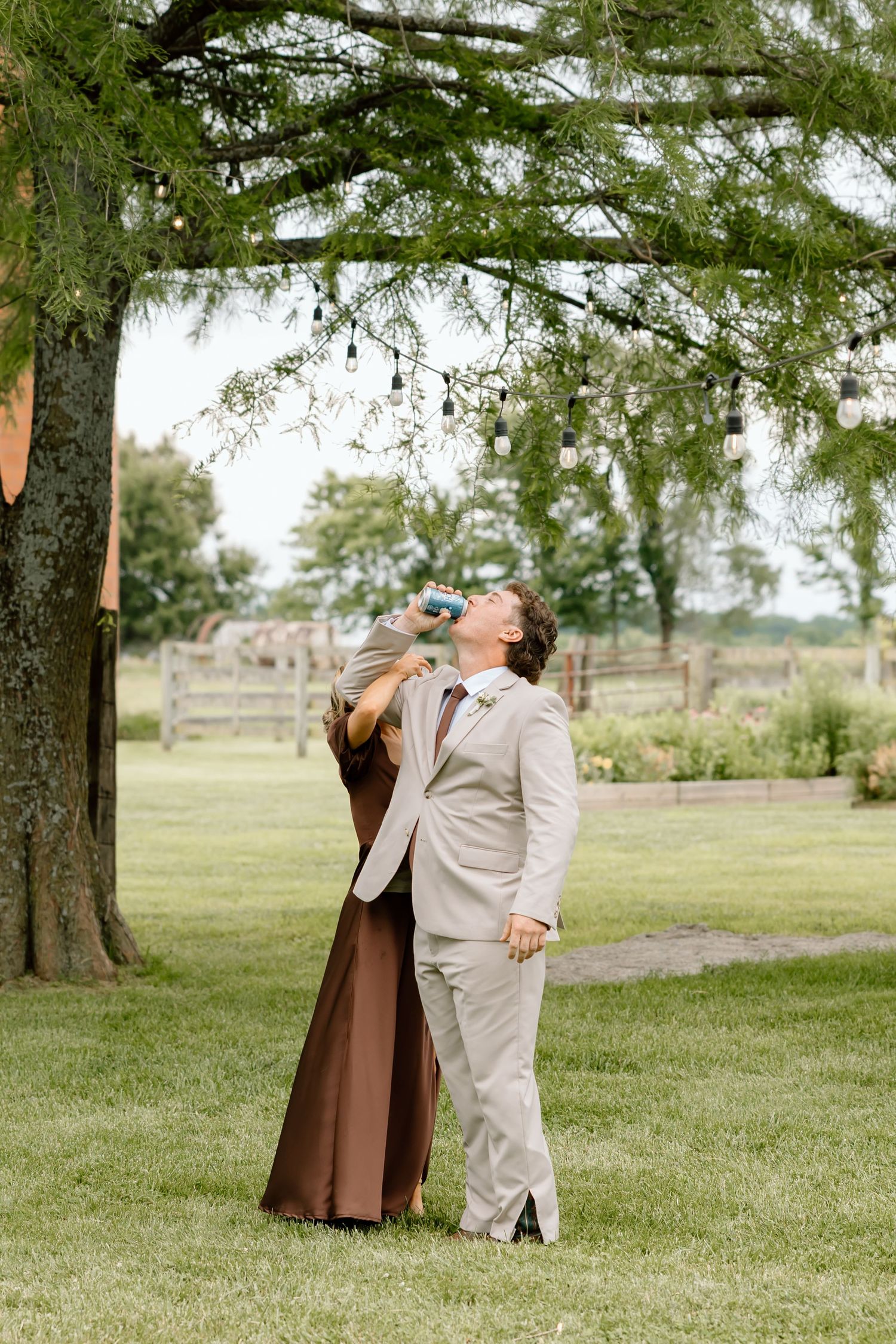 Groom in tan suit takes a drink outdoors at rustic farm wedding venue with string lights hanging from tree.