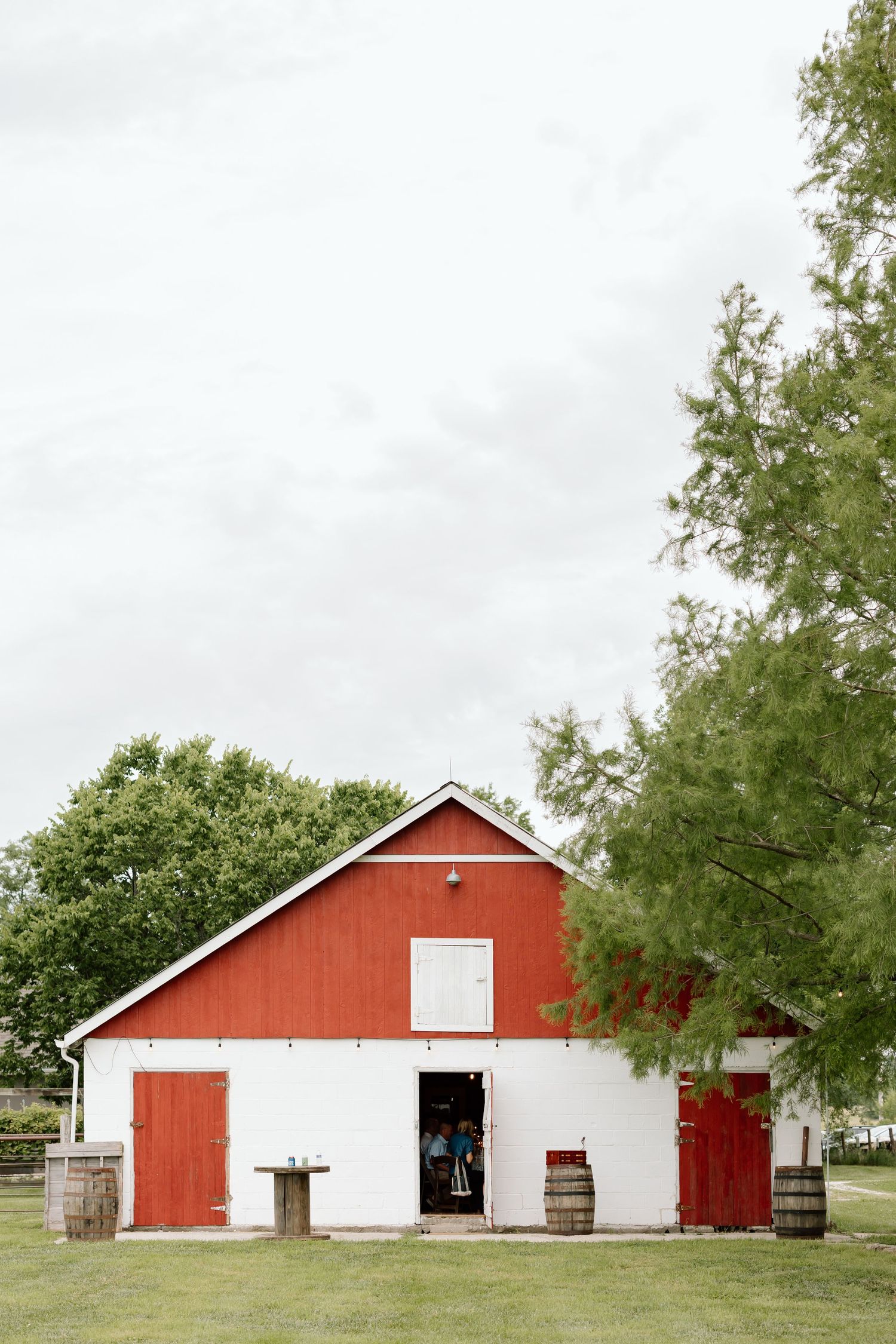 Red and white barn with wooden barrels and green trees on a cloudy day at a rustic farm venue.