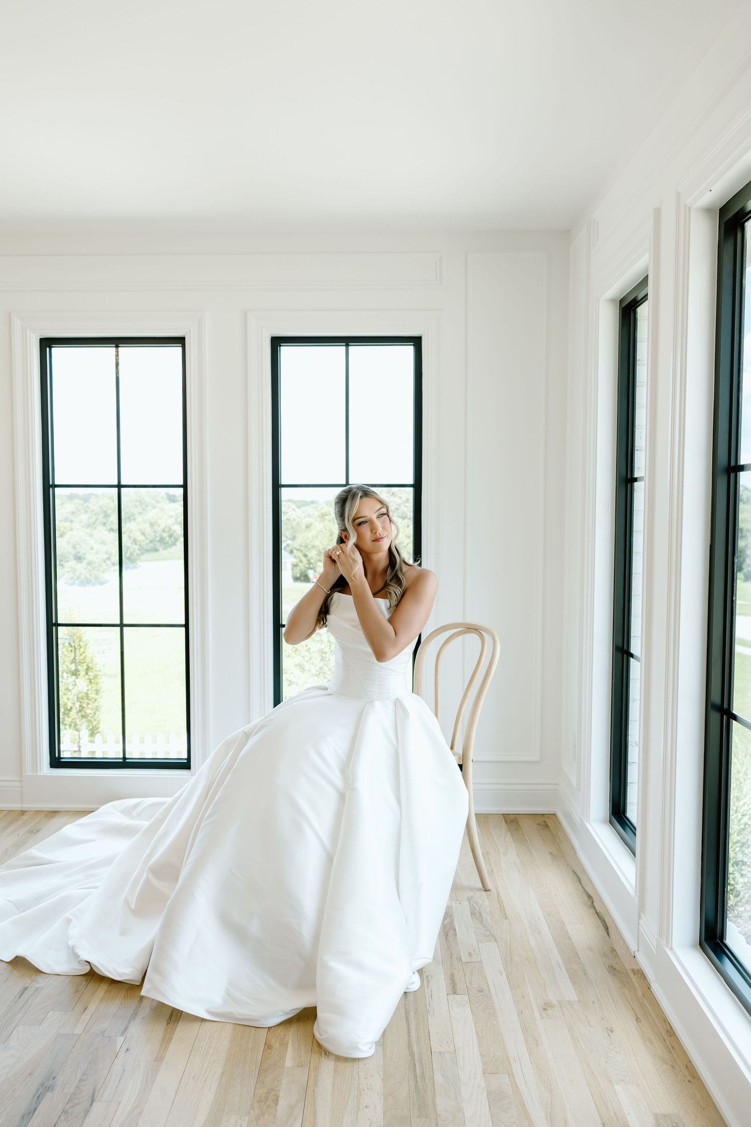 Bride in a white ballgown wedding dress seated by black-framed windows in a bright white room with hardwood floors.