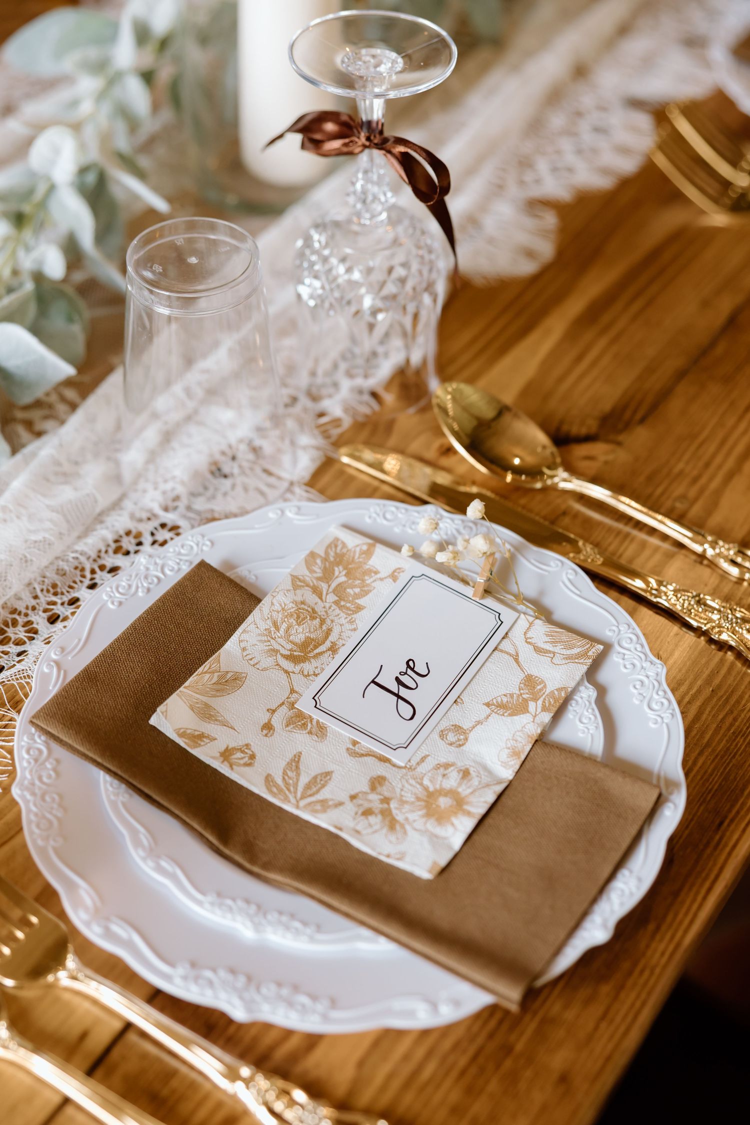 Elegant wedding table setting with gold cutlery, white lace plate, kraft napkin, floral card, and crystal glasses on wood.