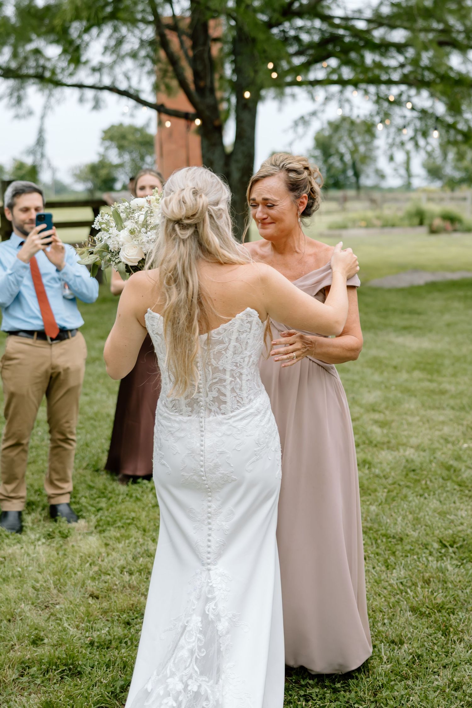 Bride in lace gown holding white bouquet shares emotional moment with woman in taupe dress at outdoor wedding ceremony.