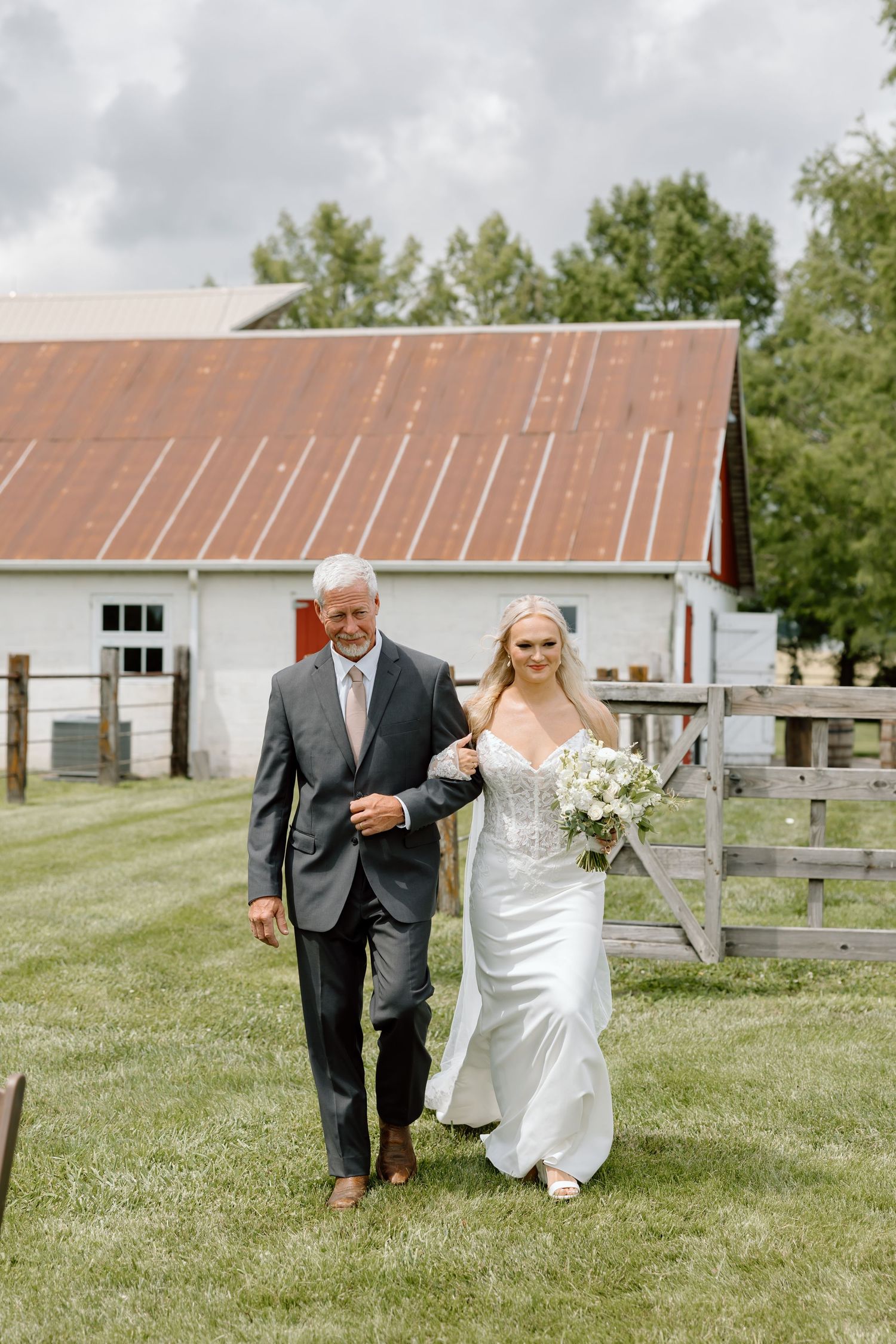 Father walks bride down outdoor aisle at rustic barn wedding venue, bride holds white bouquet in satin gown.