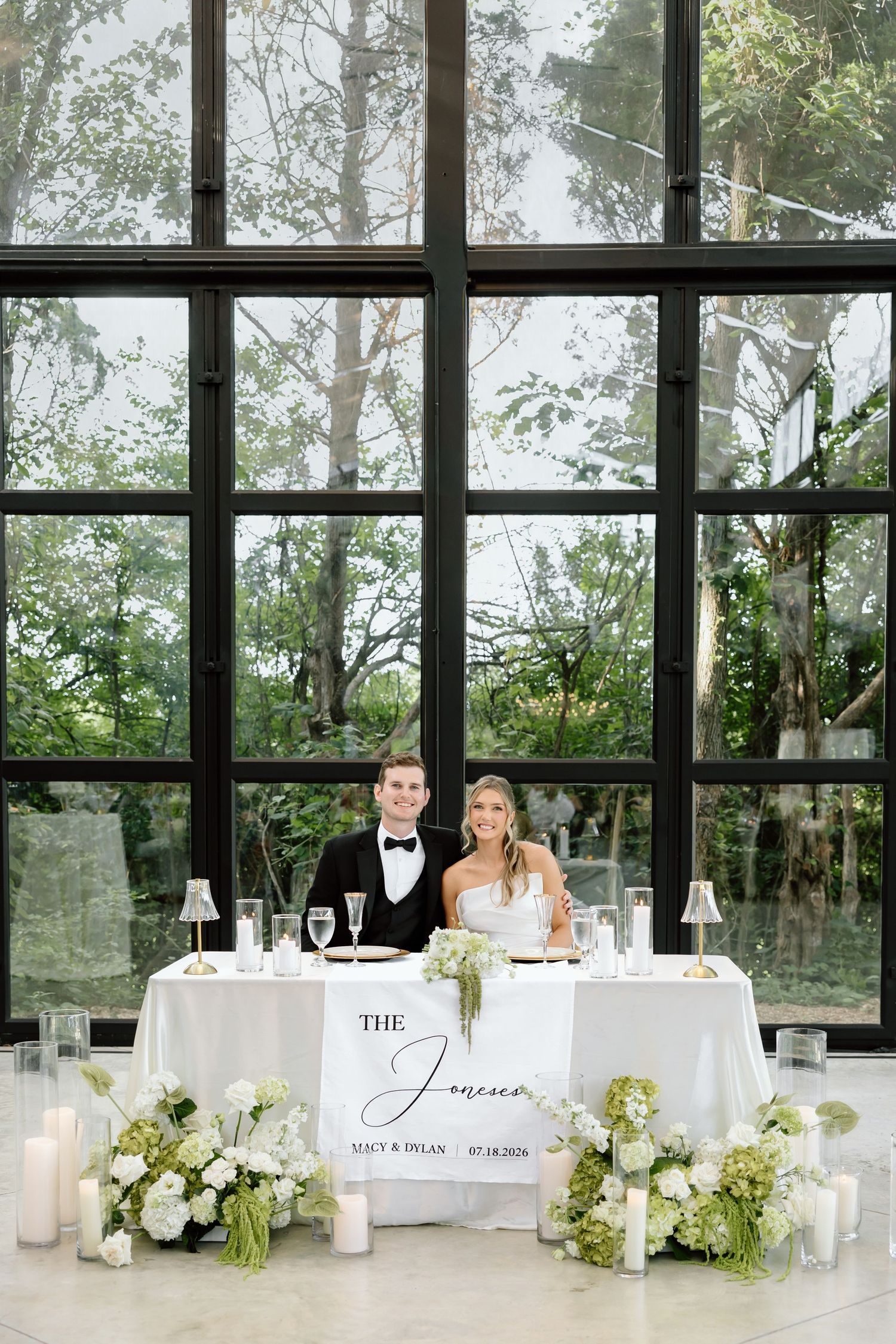Bride and groom smiling at sweetheart table with white florals and candles in front of large glass windows.