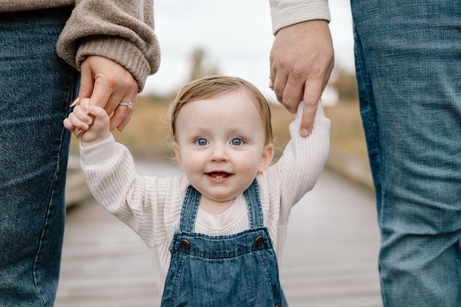A small child in overalls holds hands with adults while learning to walk on a paved surface.