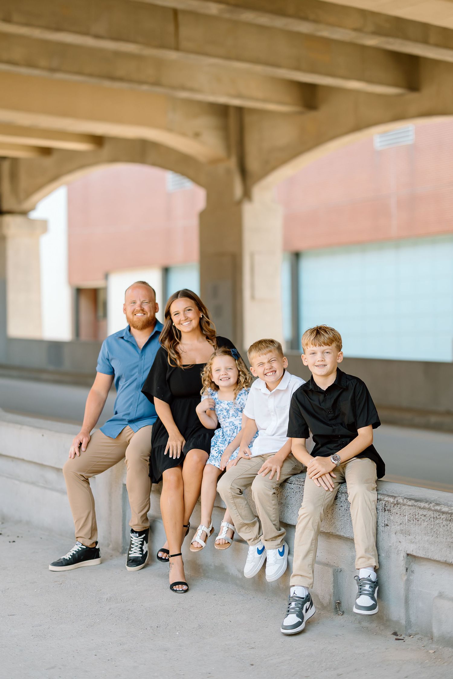 A family sits together on a concrete ledge under an urban overpass with pink walls in the background.