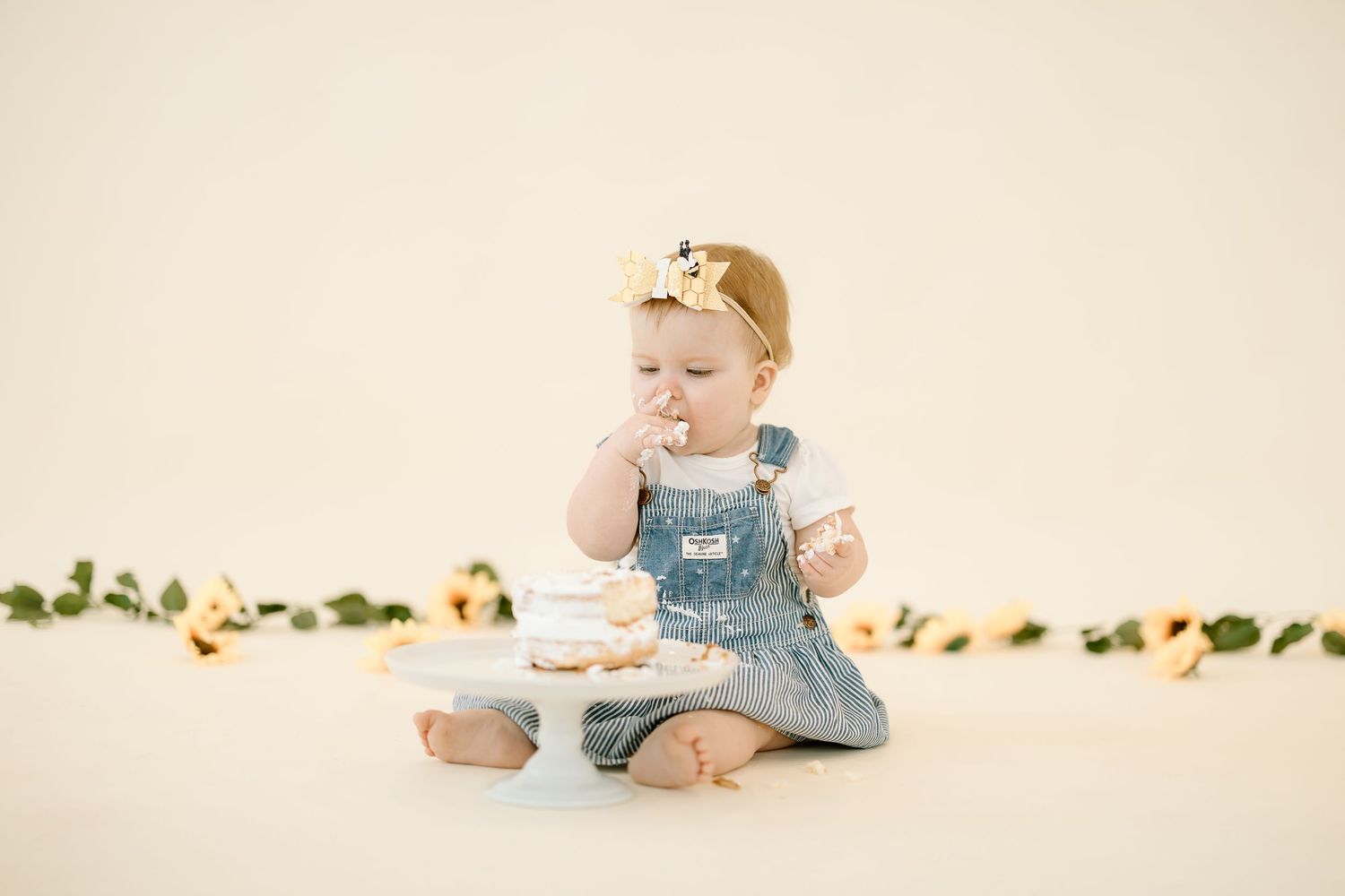 Baby in blue denim overalls sits on cream colored backdrop with decorative greenery garland.