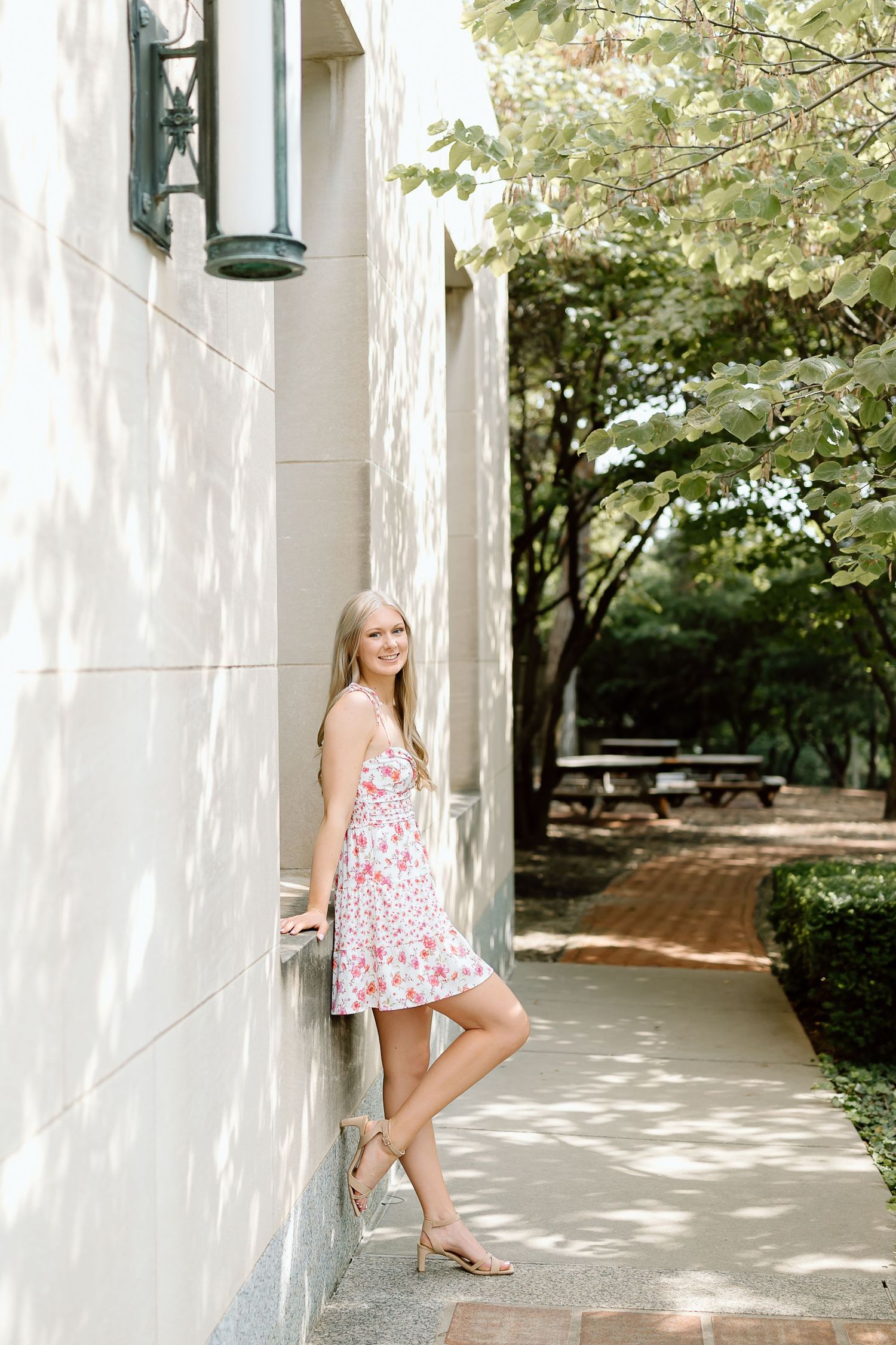 A summery floral dress stands out against a white building wall along a tree-lined walkway on a bright day.