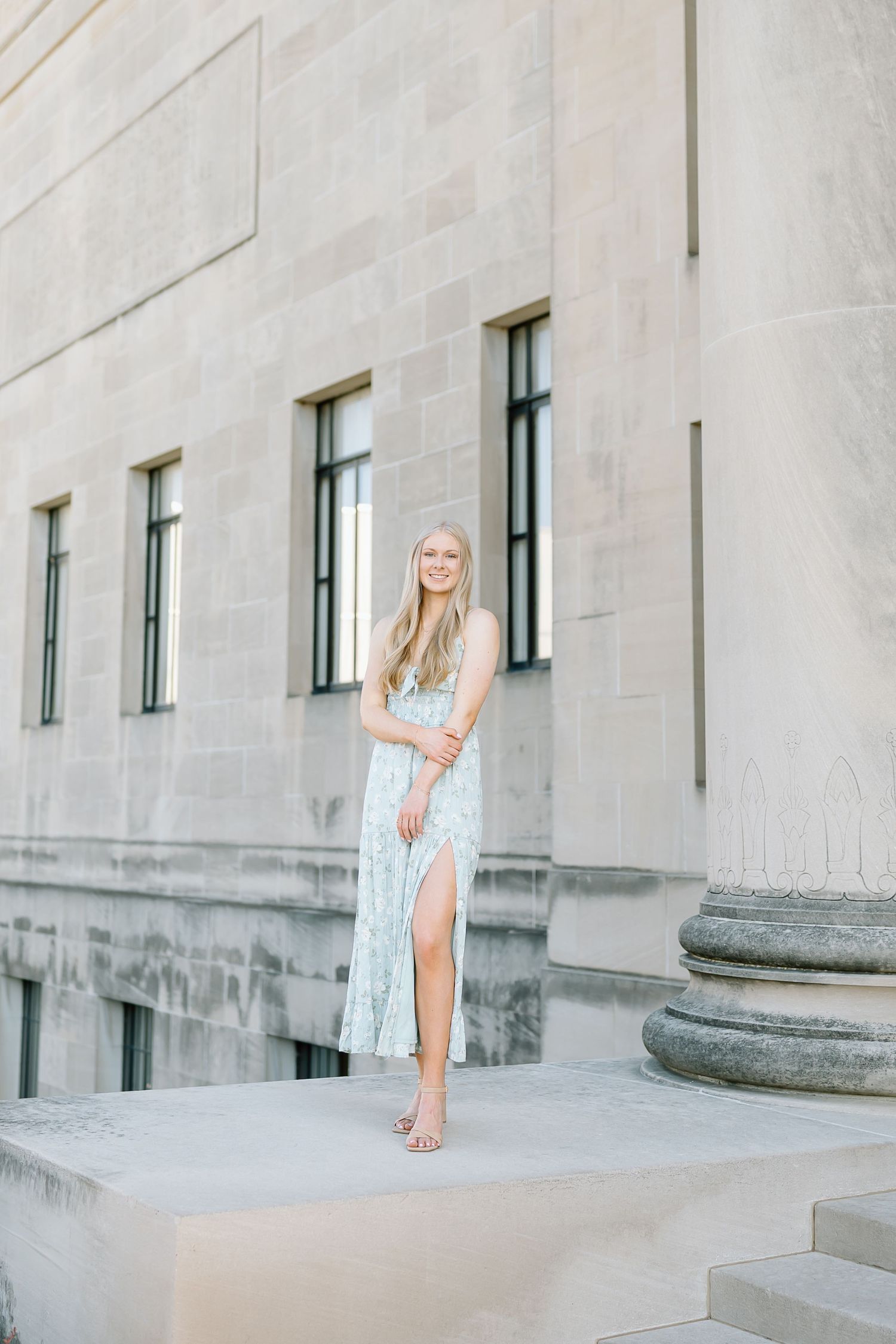 A person in a white dress poses between stone columns on the steps of a grand building.