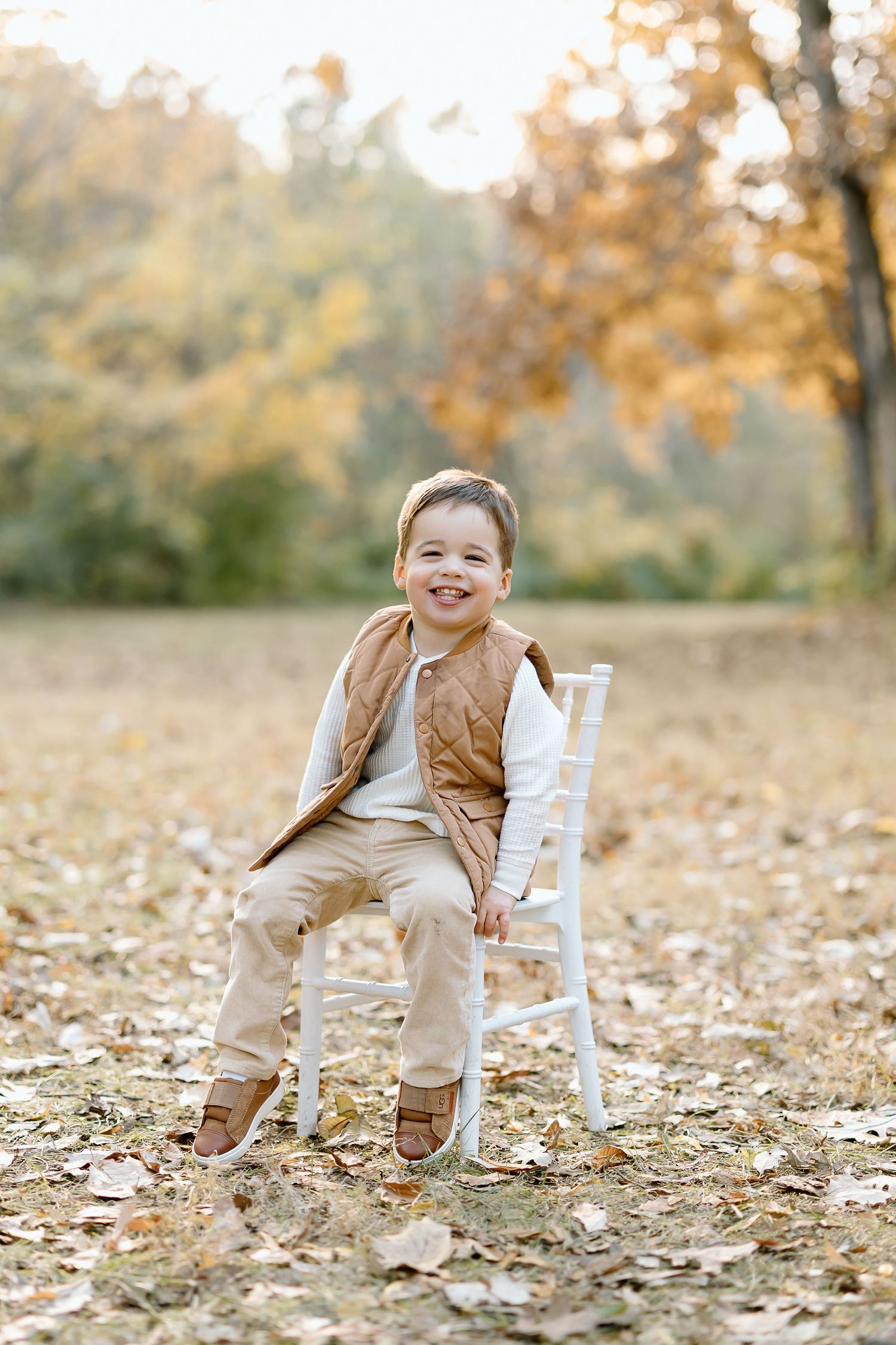 A child sits on a white wooden chair surrounded by fallen autumn leaves in a park setting.