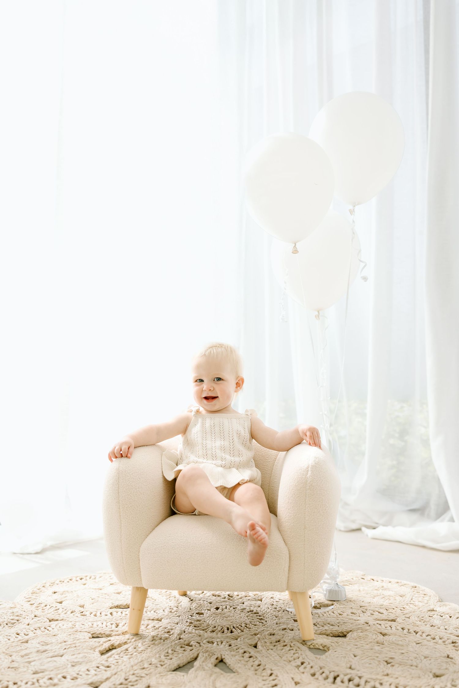 Happy baby sitting in a white chair against a bright window with sheer curtains and a patterned rug.