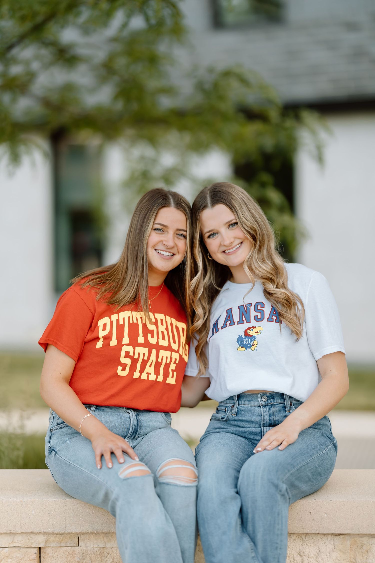 Two friends wearing college sweatshirts and ripped jeans sit together on a stone wall outdoors.