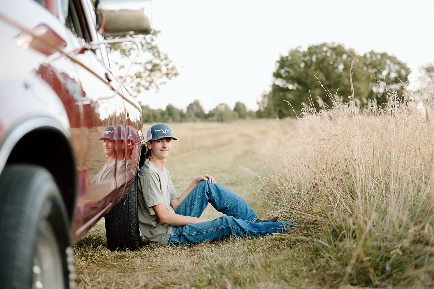 A person in casual attire sits relaxed in tall grass near an old red truck in a rustic outdoor setting.
