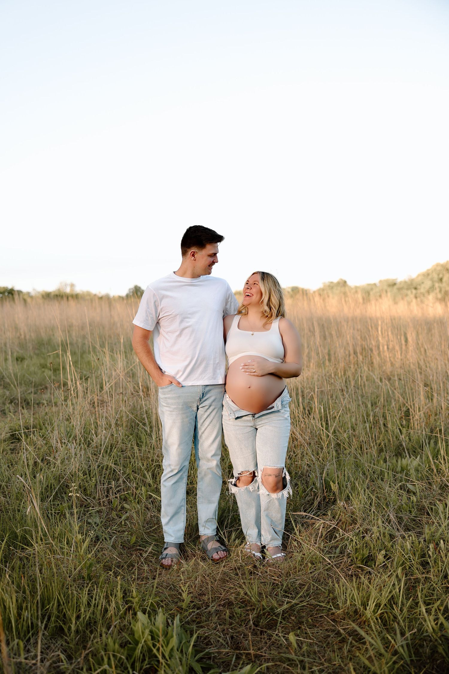 A couple and small child stand together in a golden sunlit meadow at sunset wearing casual white and denim outfits.