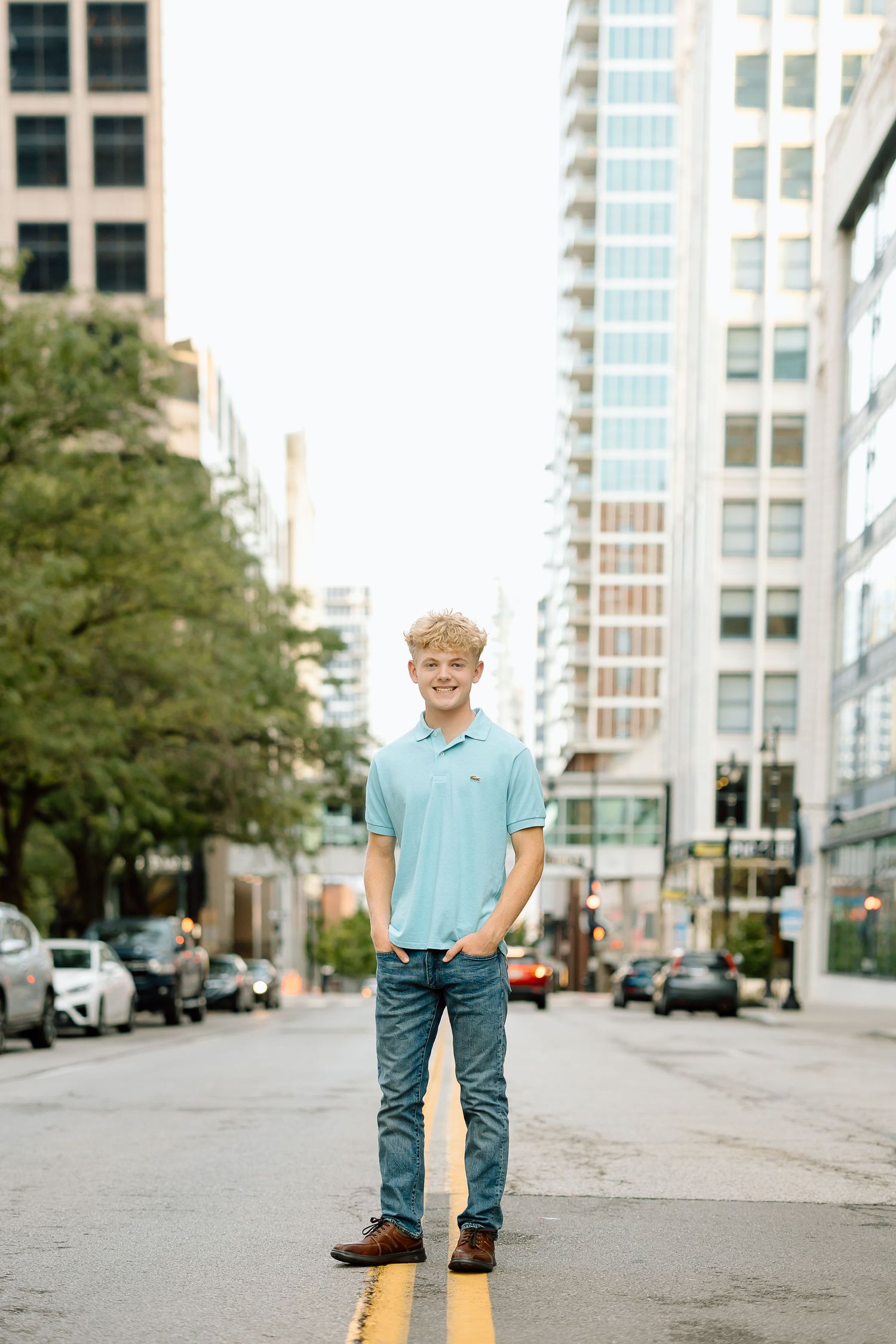 Person in light blue shirt and jeans stands on an empty urban street with tall buildings in the background.