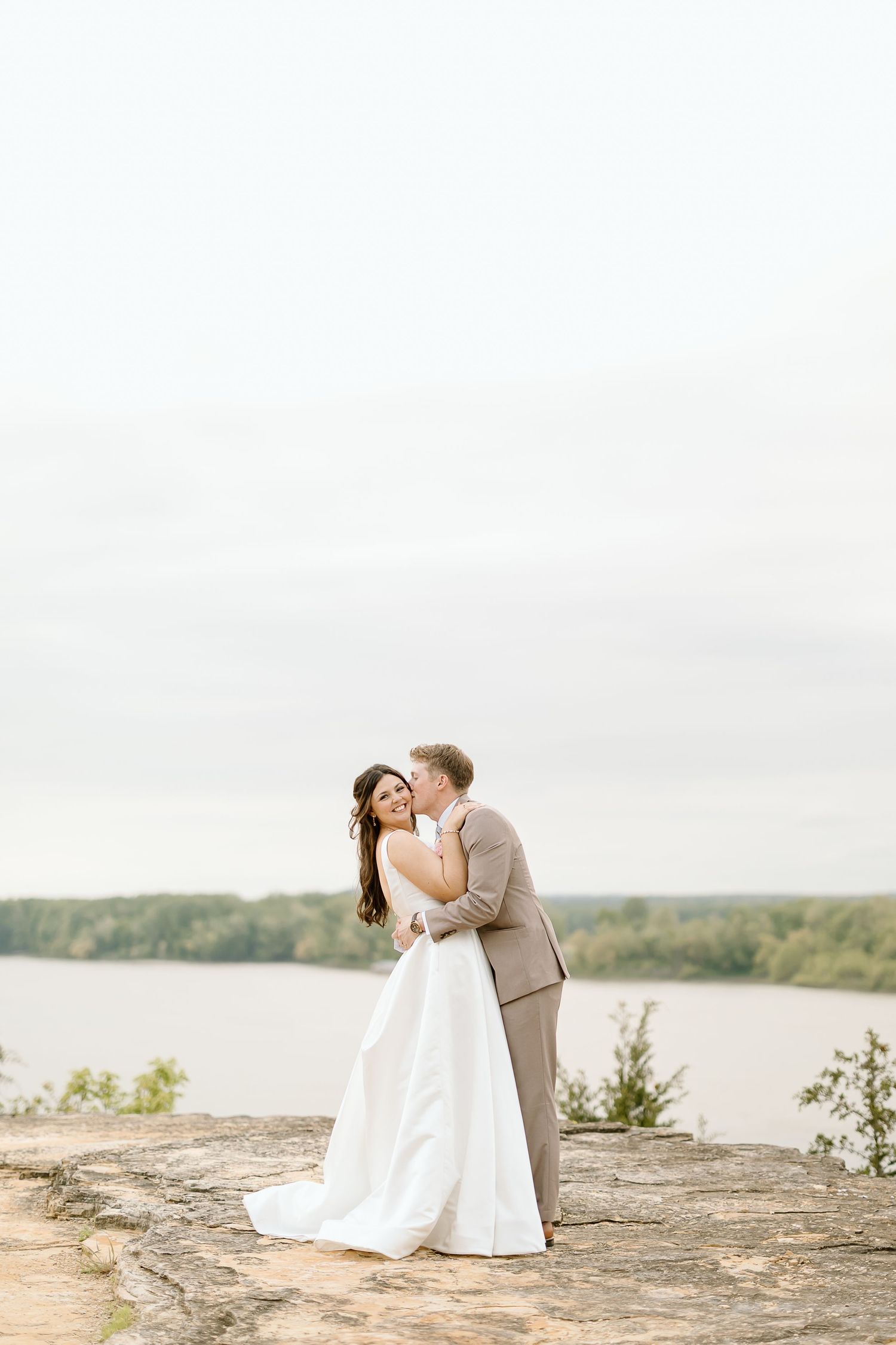 A romantic couple in wedding attire embrace on a scenic cliff overlooking a serene lake during sunset.