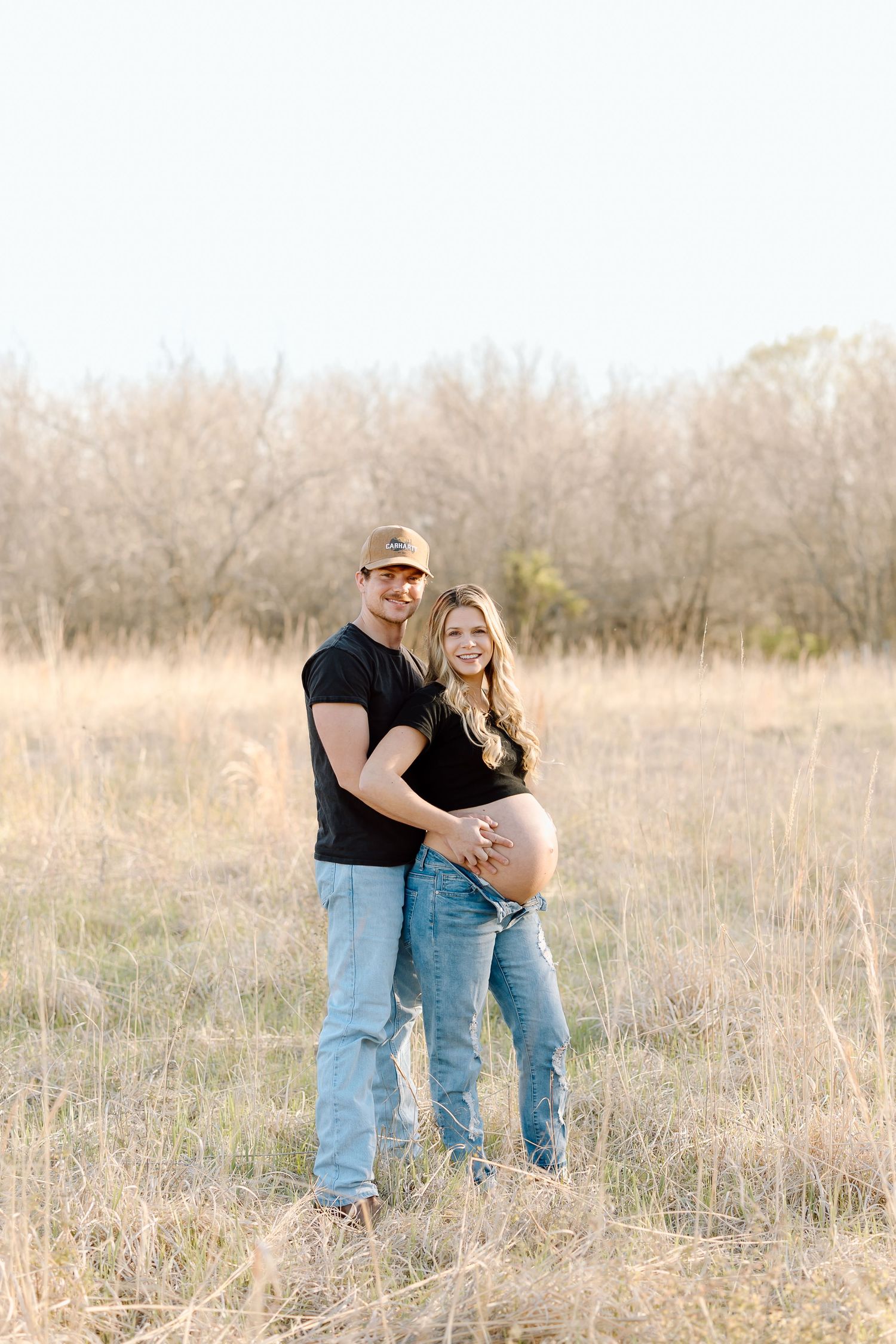 A couple stands together in a golden field during sunset, embracing and showing off her pregnant belly.