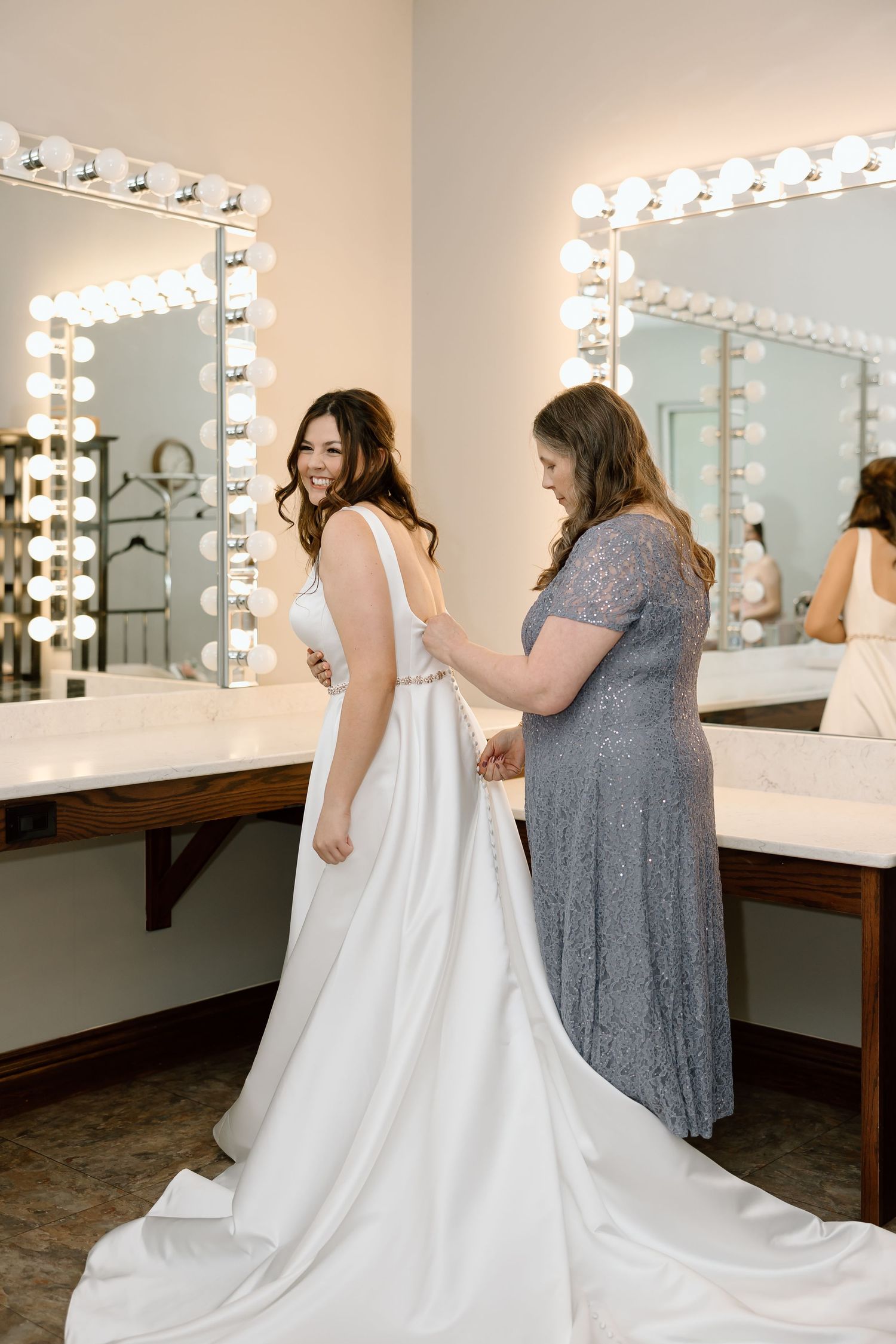 A wedding dress fitting scene in a bridal dressing room with illuminated mirrors and helping hands adjusting the gown.