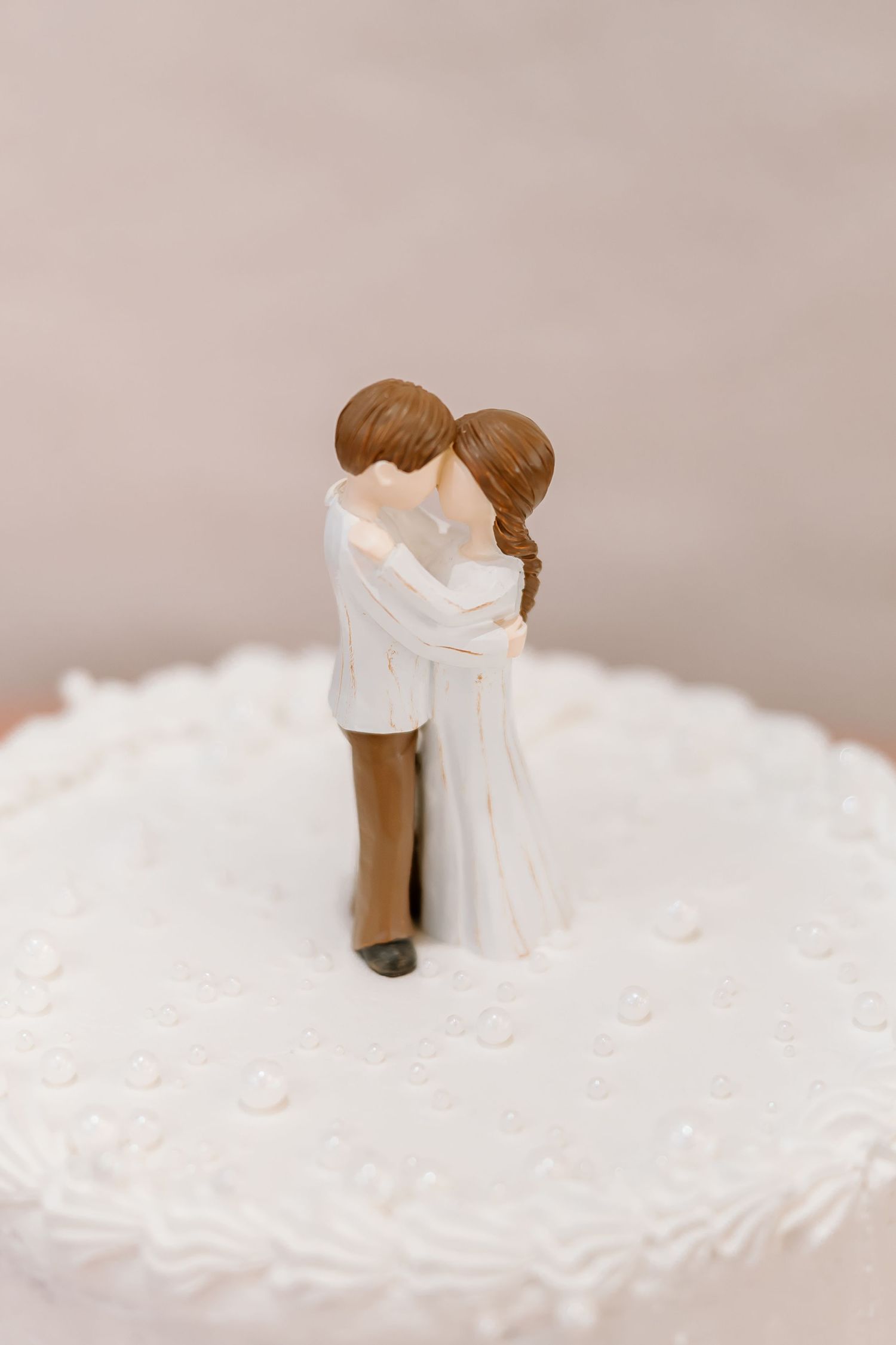 A wedding cake topper shows a romantic couple in formal attire embracing against a soft gray background.