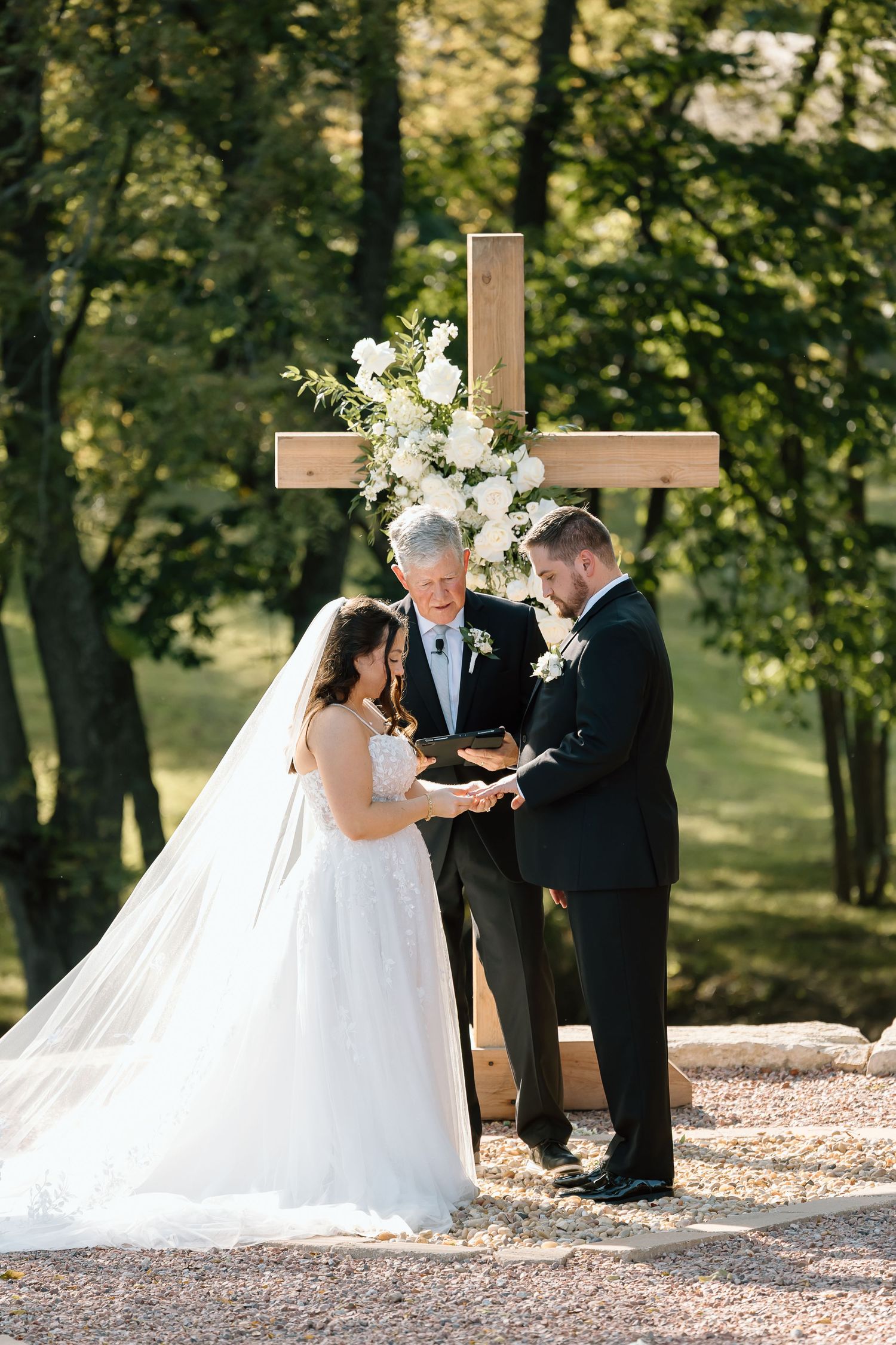 Outdoor wedding ceremony taking place on a gravel path surrounded by trees with white floral decorations.