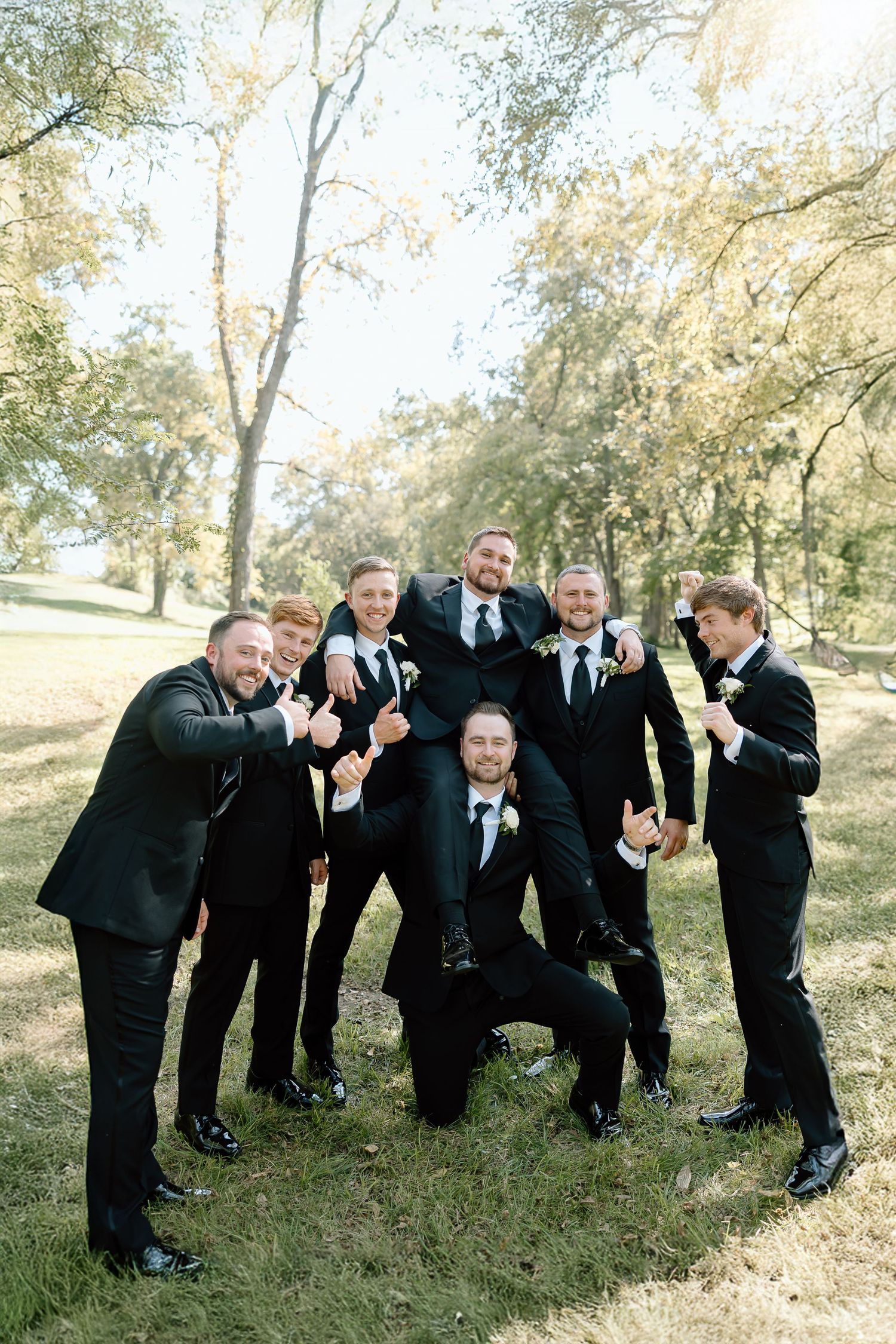 Groomsmen in black suits perform a domino effect falling sequence outdoors in a grassy area on a sunny day.